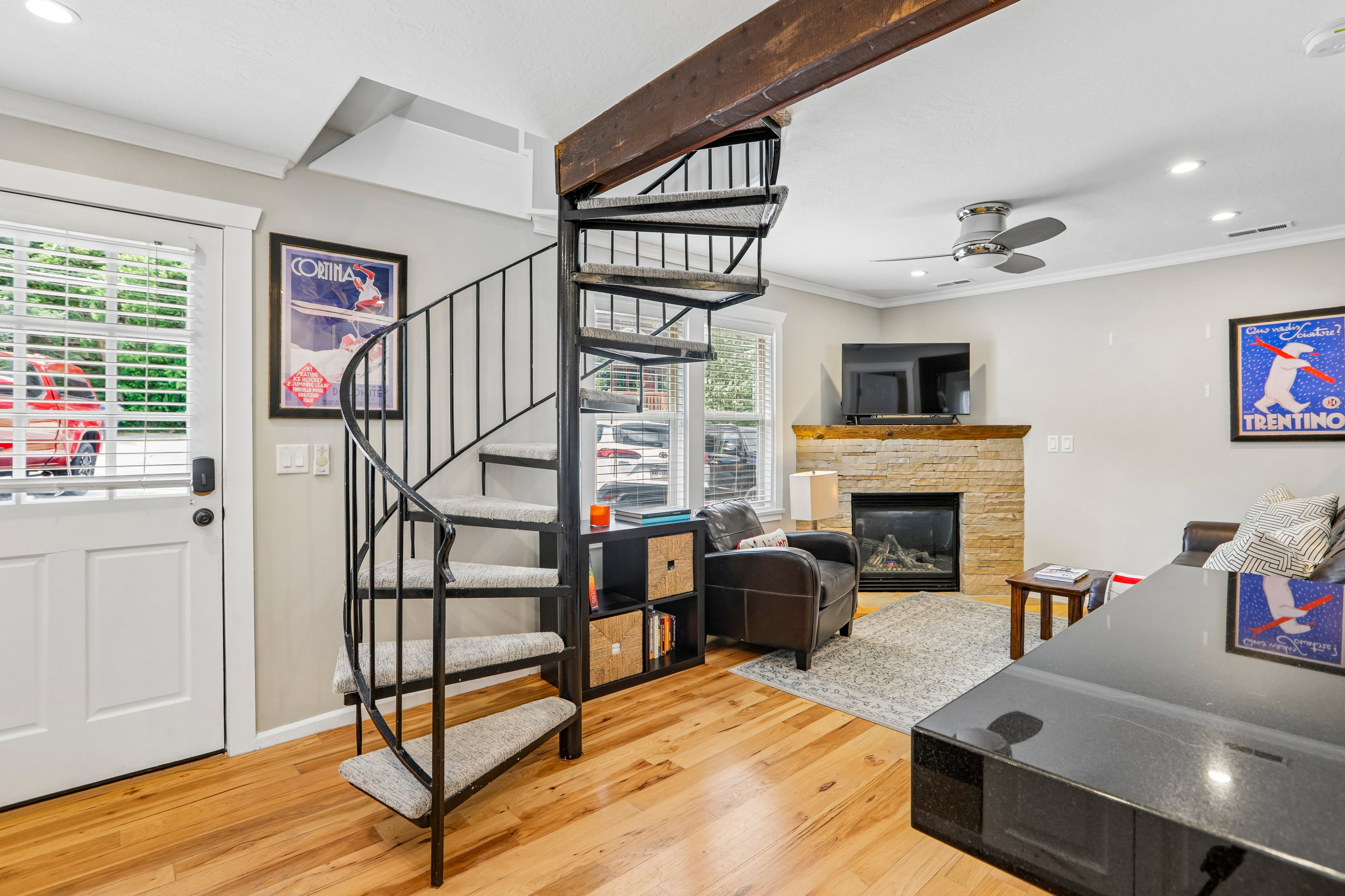 Spiral staircase with metal railing and carpeted treads, wood-finish flooring, stone-faced fireplace with wood mantle, overhead ceiling fan, and recessed lighting