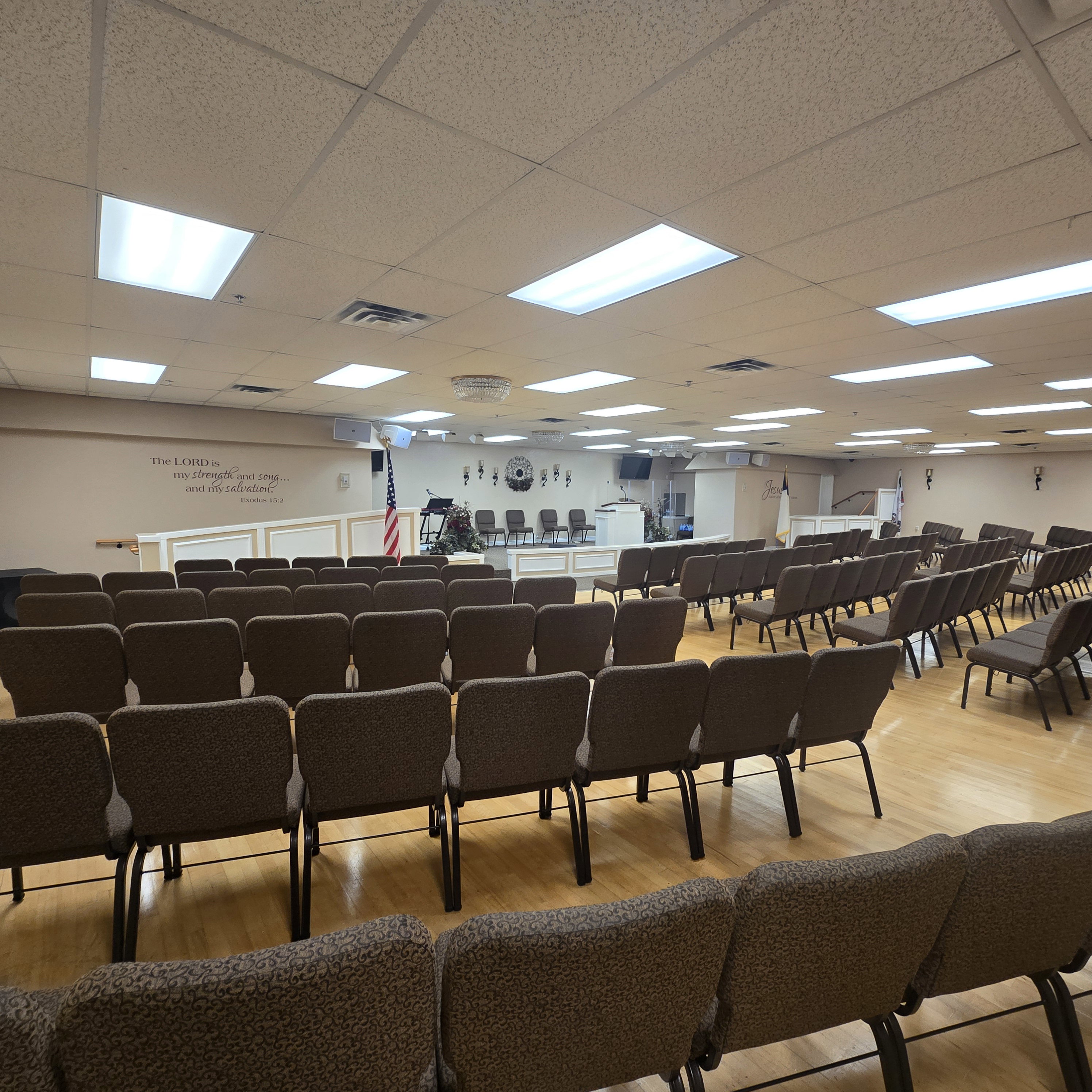 Spacious assembly hall featuring wood-finish flooring, drop ceiling with integrated lighting, and a raised platform stage