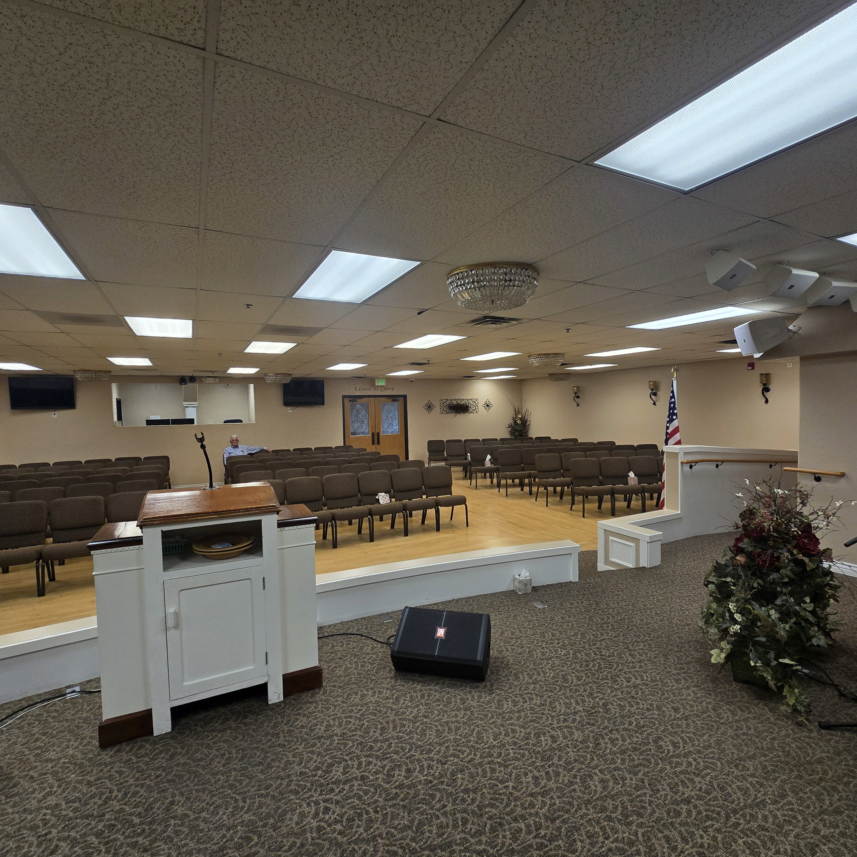 Spacious assembly room featuring a raised platform with wood-finish flooring, recessed fluorescent lighting, and a textured drop ceiling