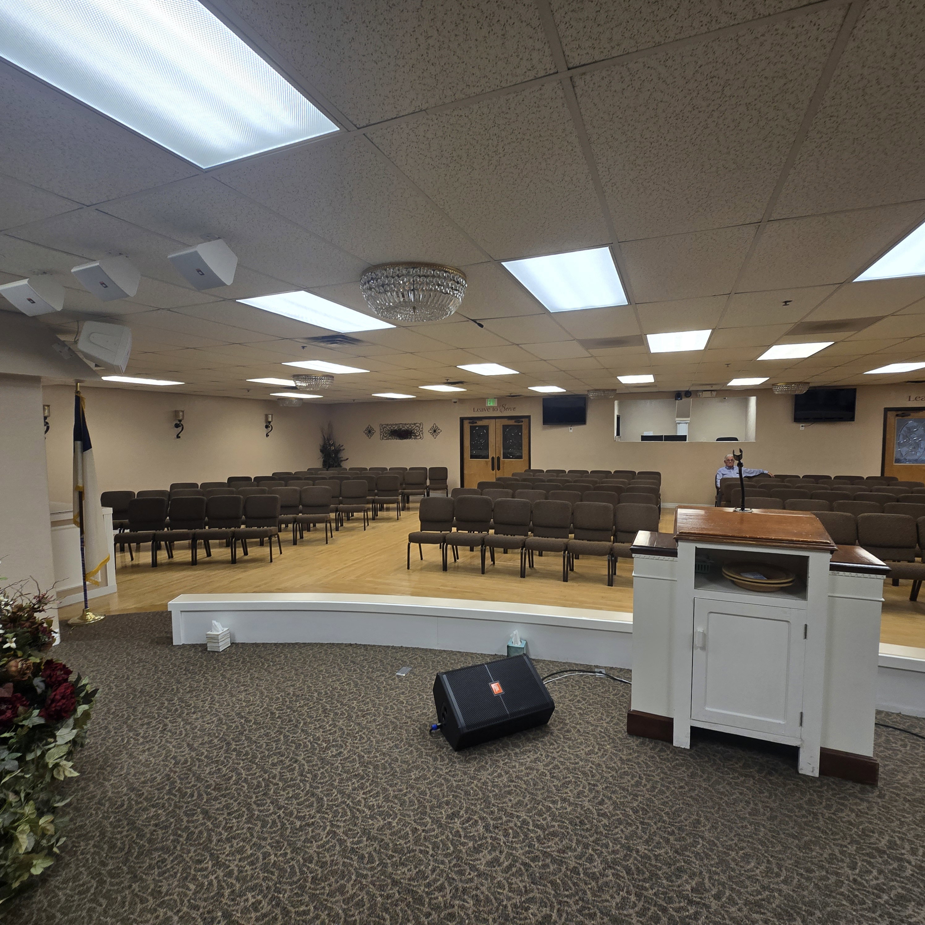 Spacious assembly room featuring a raised stage with wood-finish flooring, recessed fluorescent lighting, and a decorative central chandelier