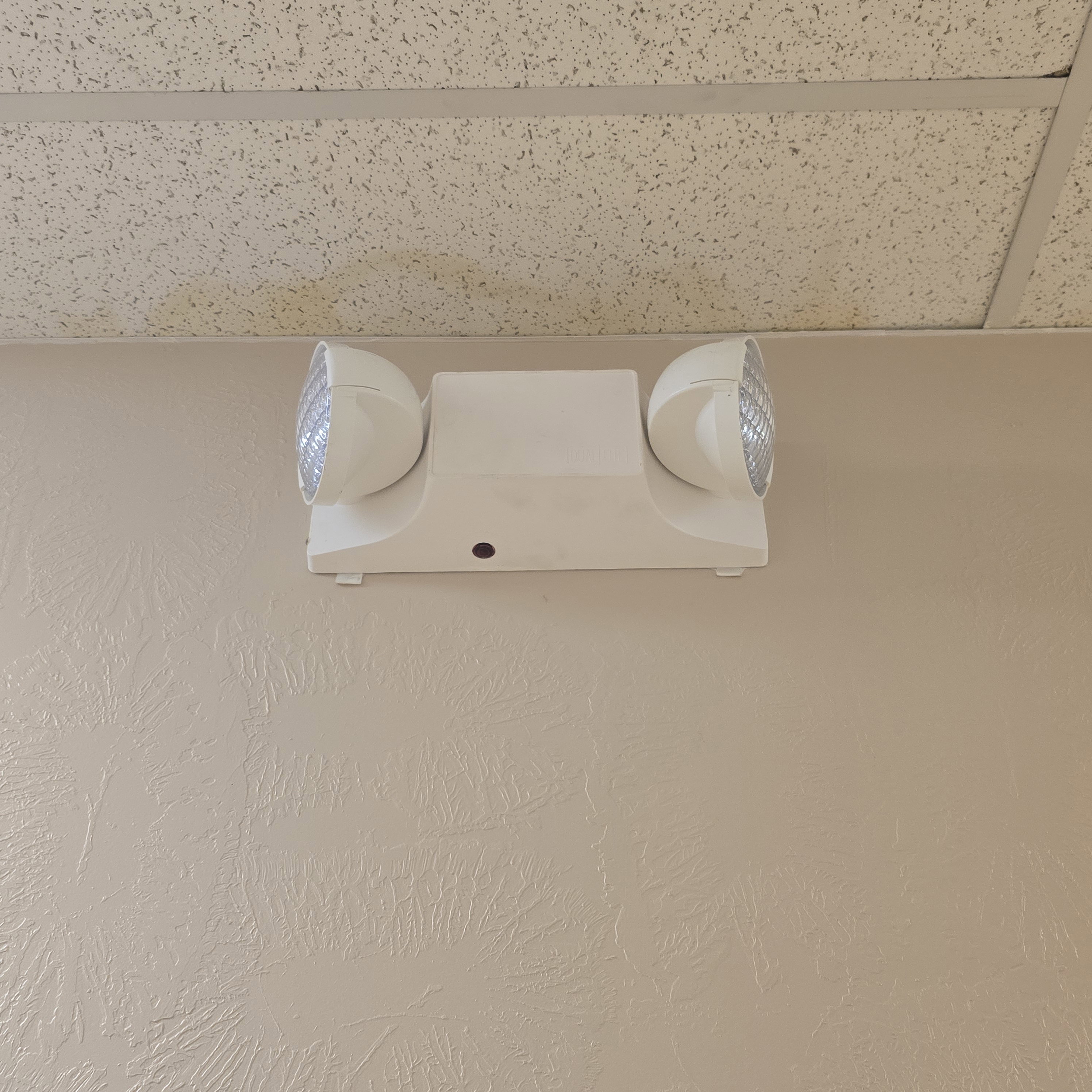 Wall-mounted emergency lighting fixture with dual adjustable heads, positioned below a suspended acoustic tile ceiling