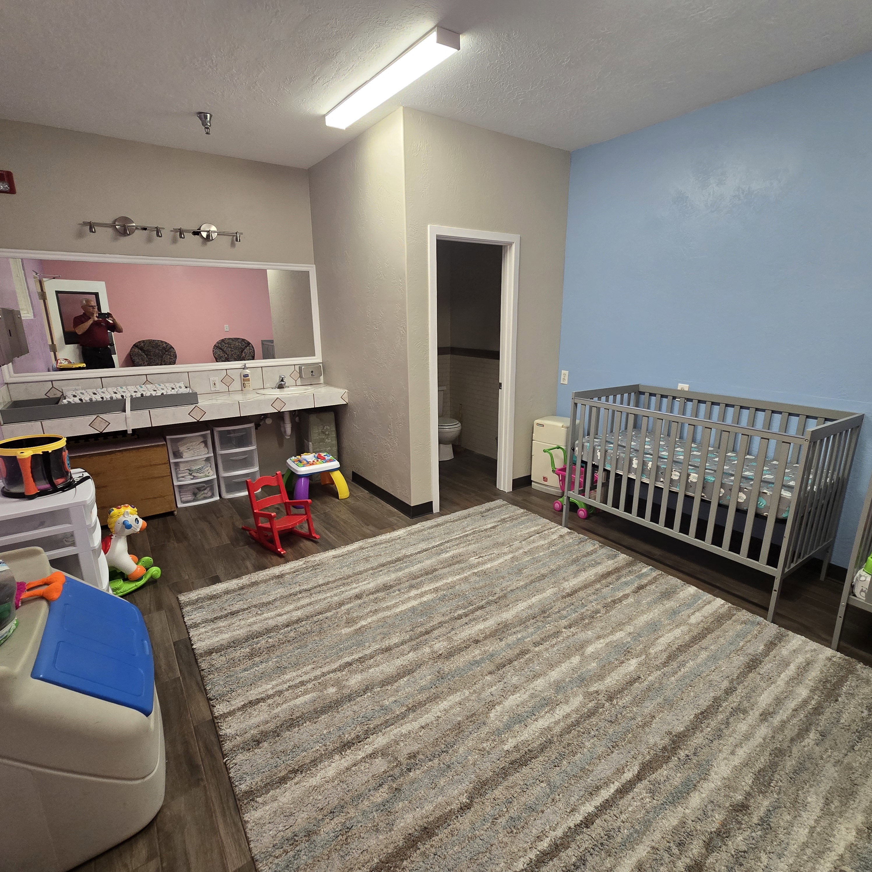 Spacious nursery room featuring wood-finish flooring, a large area rug, and dual vanity stations with tiled countertops. Toilet in closed area.