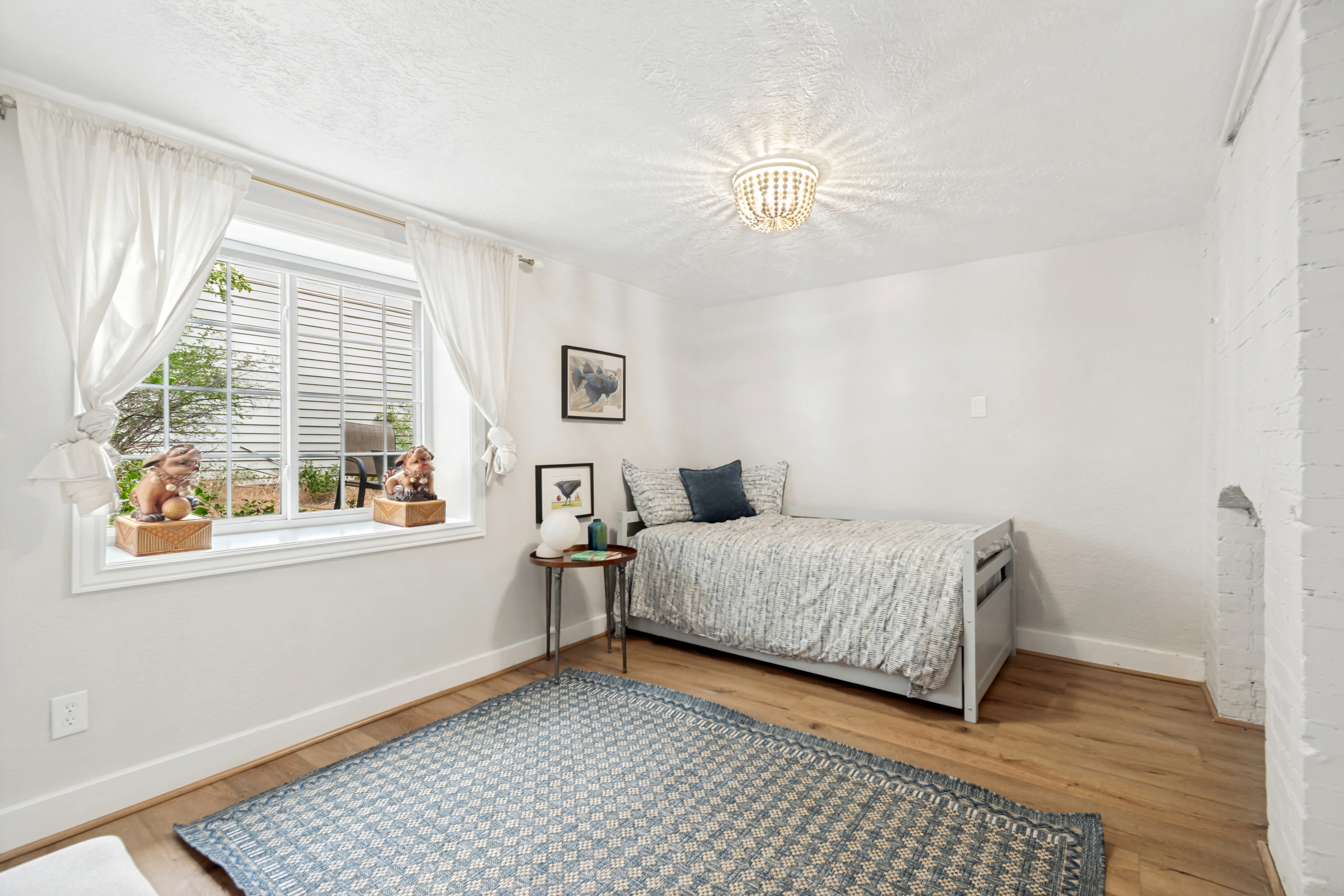 Room featuring a large window with privacy blinds, wood-finish flooring, a white-painted brick wall, and a beaded chandelier