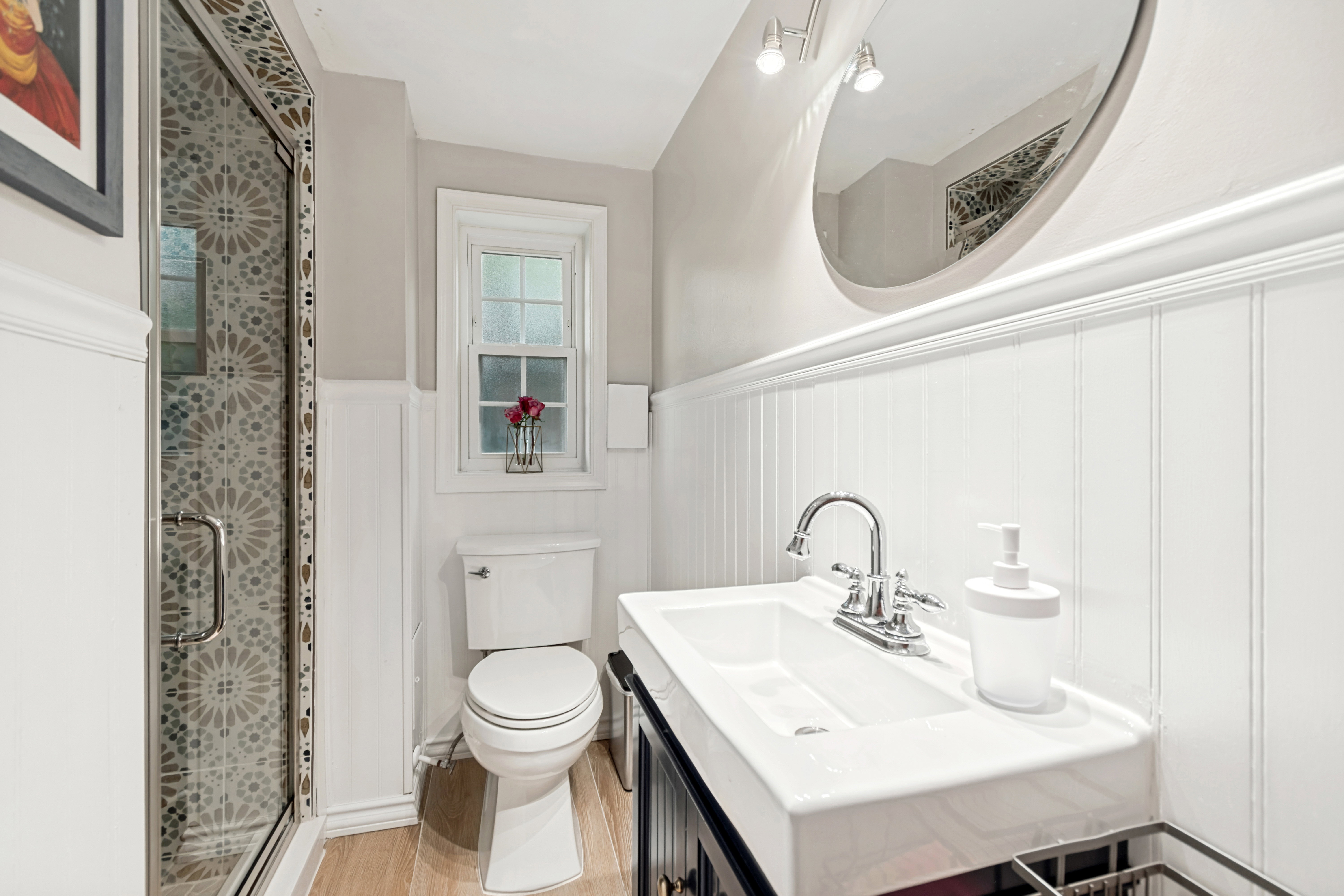 Bathroom featuring a glass-enclosed shower with patterned tile, beadboard wainscoting, a rectangular sink with chrome fixtures, a dark wood-finish vanity, and a window with white trim