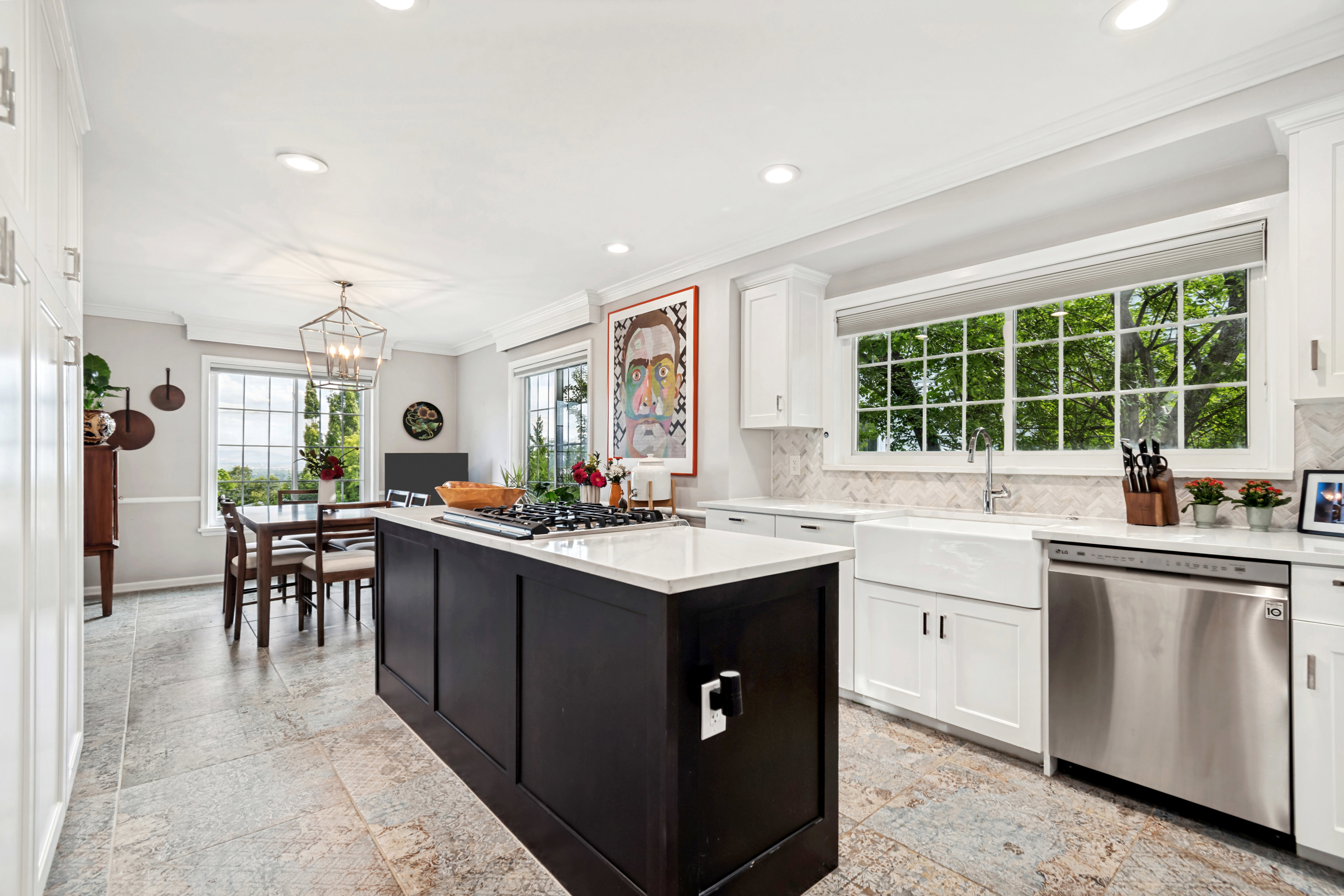 Spacious kitchen featuring a dark island with cooktop, white cabinetry, a farmhouse sink, stainless steel appliances, and large windows with transom details