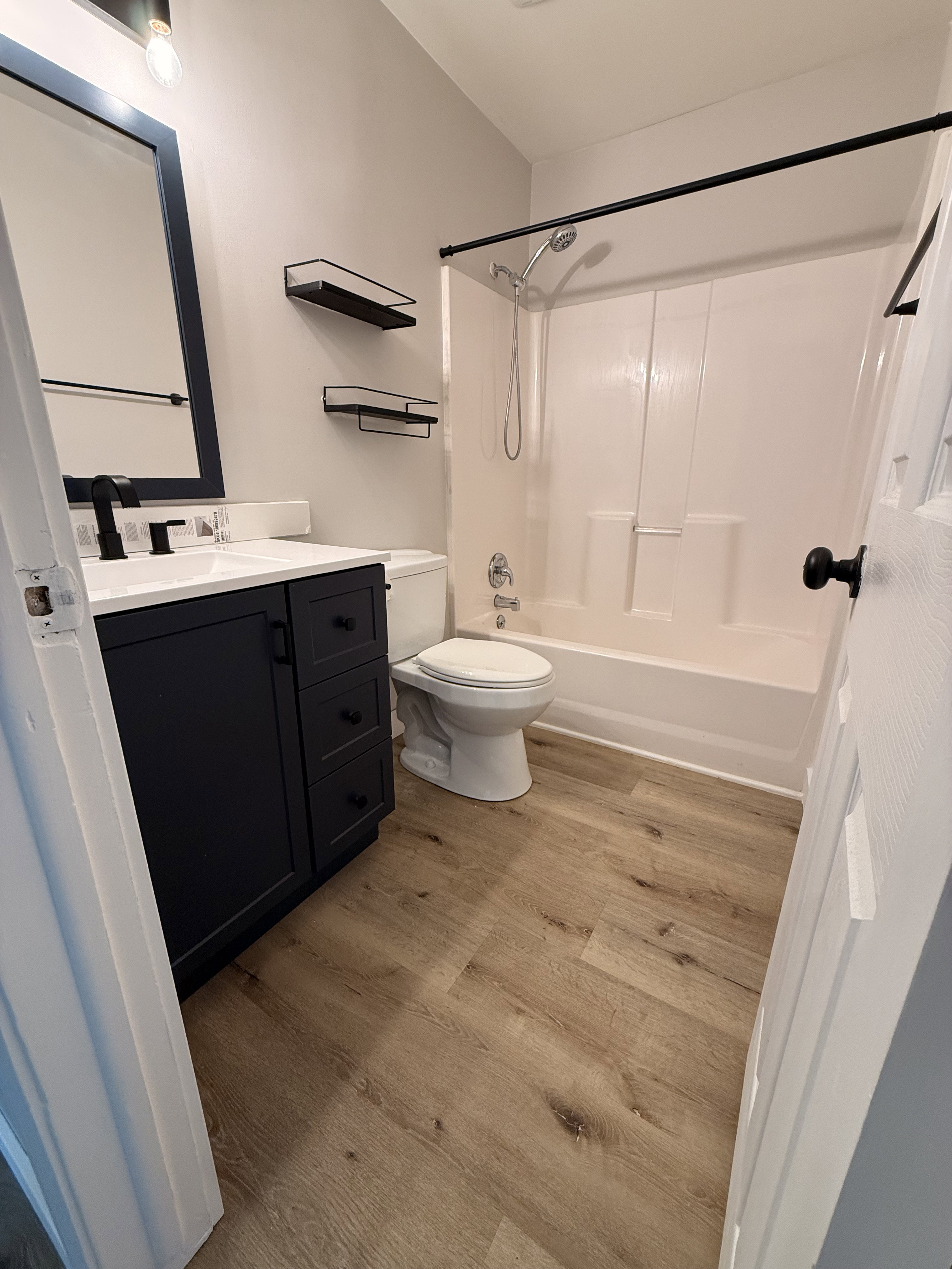 Bathroom featuring a dark wood-finish vanity with white countertop, black framed mirror, wood-finish flooring, white tub with shower, and matte black fixtures