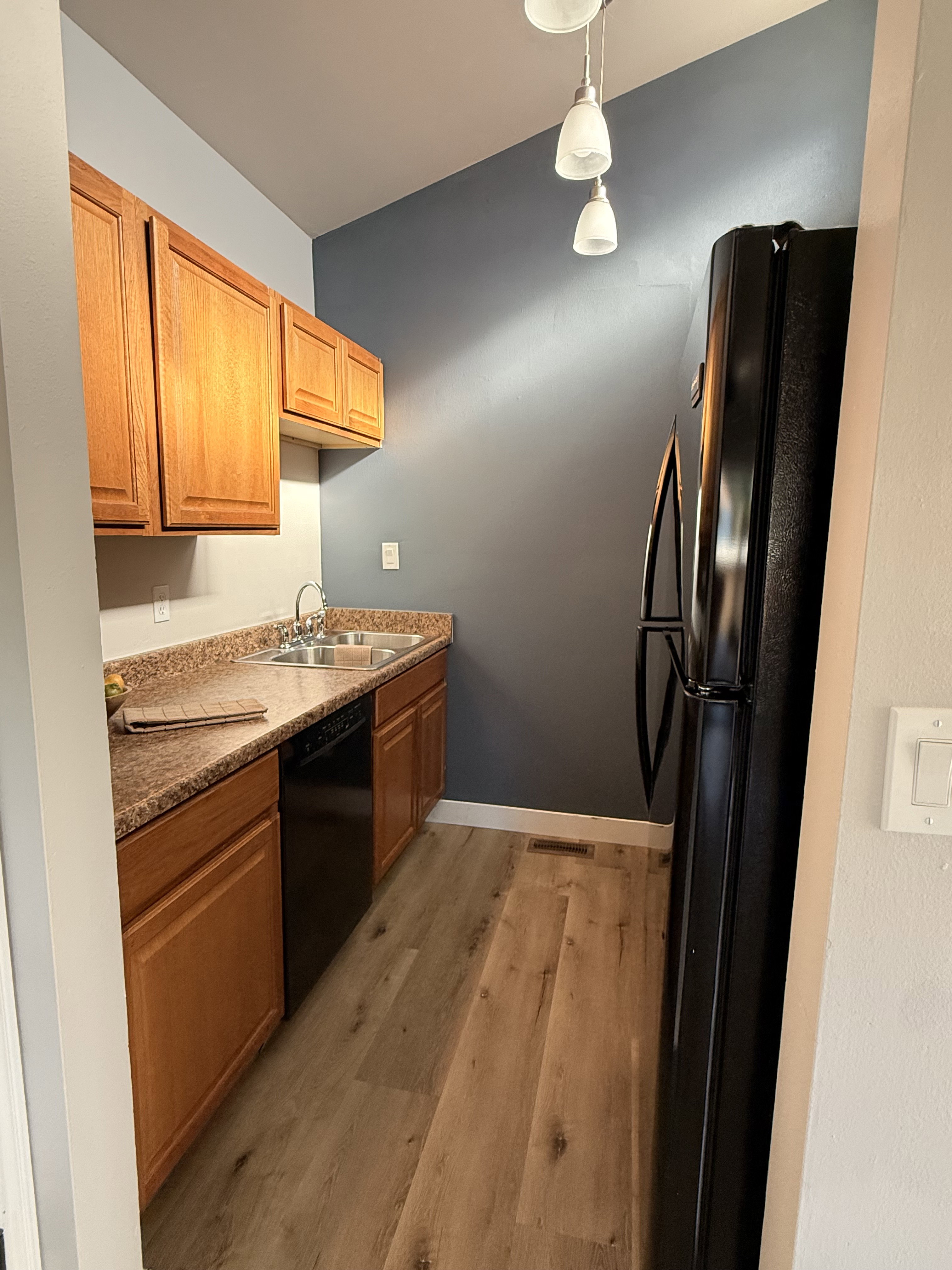 Kitchen featuring wood-finish flooring, oak-toned cabinetry, a double basin sink, black appliances, and pendant lighting