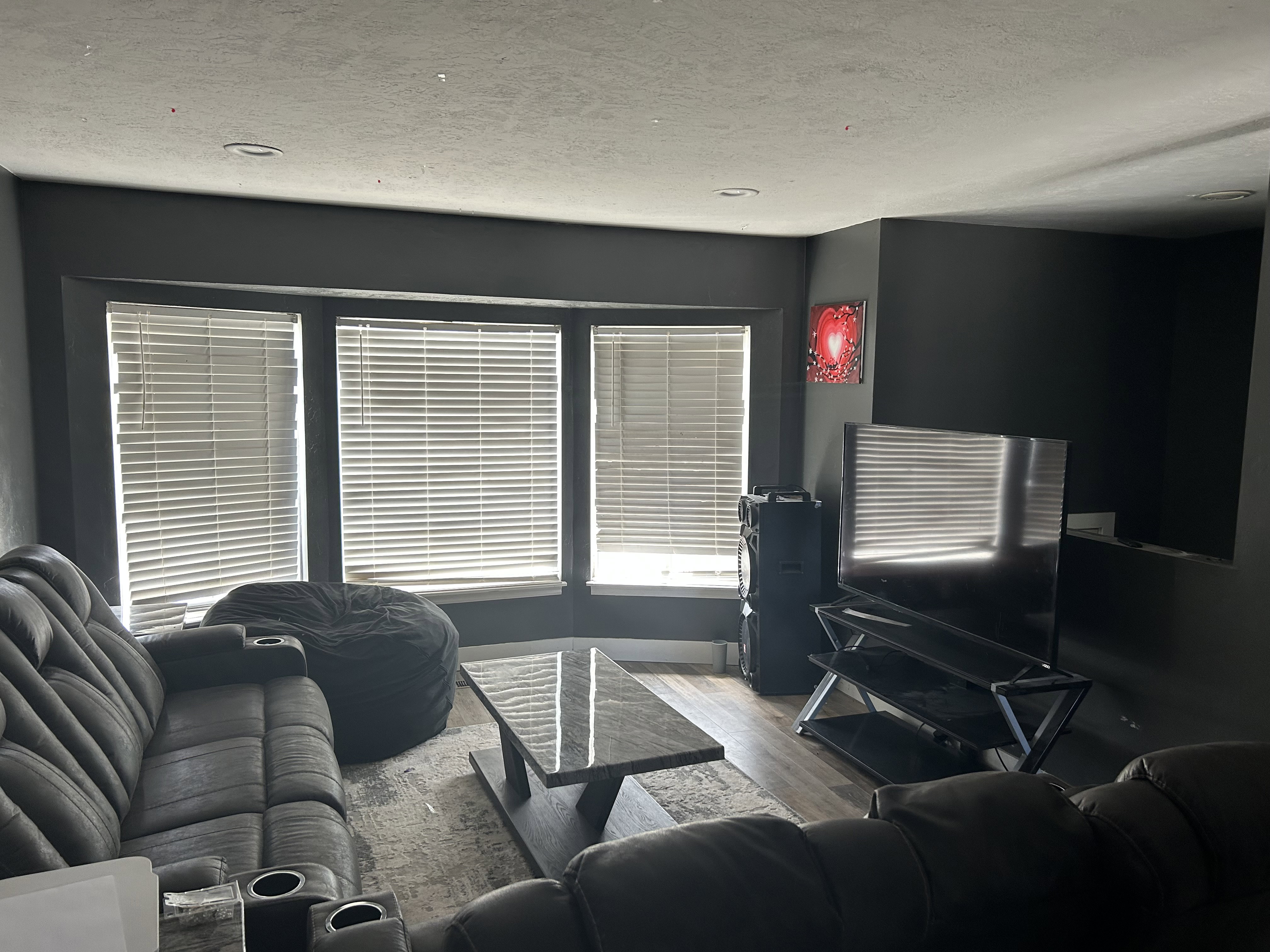 Living area featuring wood-finish flooring, a large bay window with horizontal blinds, recessed ceiling lighting, and dark-toned wall finishes