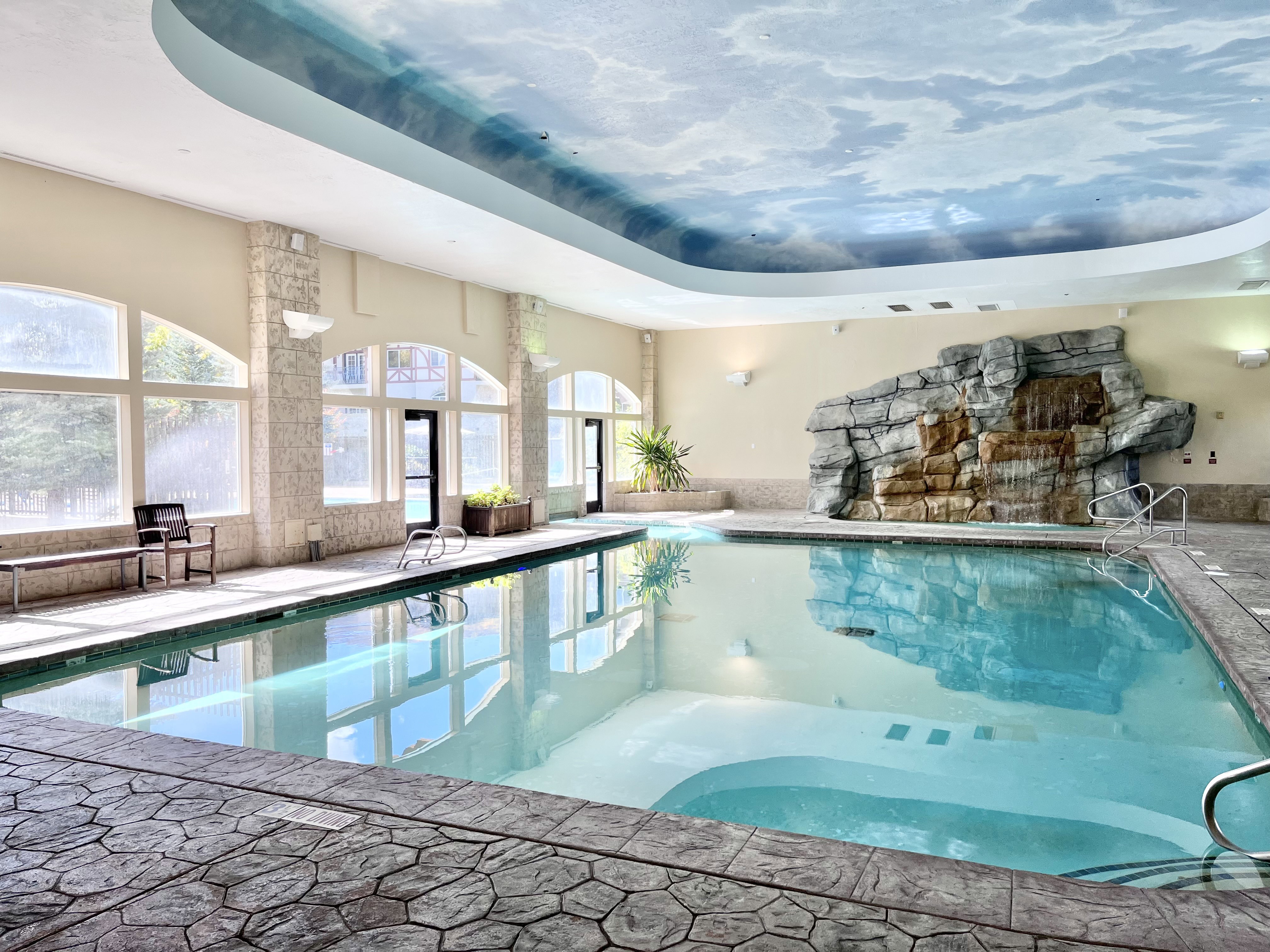 Indoor swimming pool featuring a waterfall rock formation and a custom cloud-mural ceiling