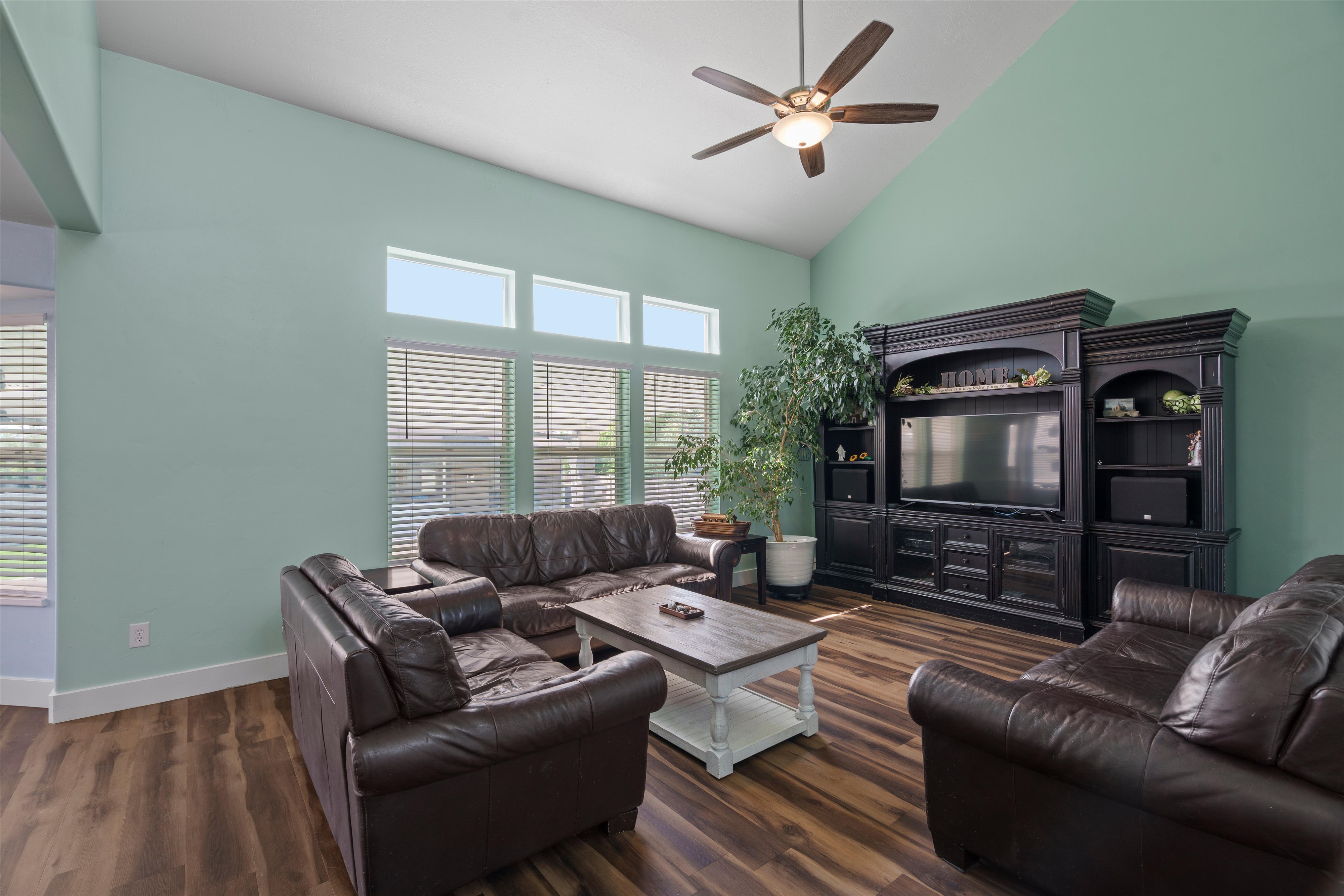 Vaulted ceiling living area featuring wood-finish flooring, a contemporary ceiling fan with integrated lighting, and multiple windows with horizontal blinds
