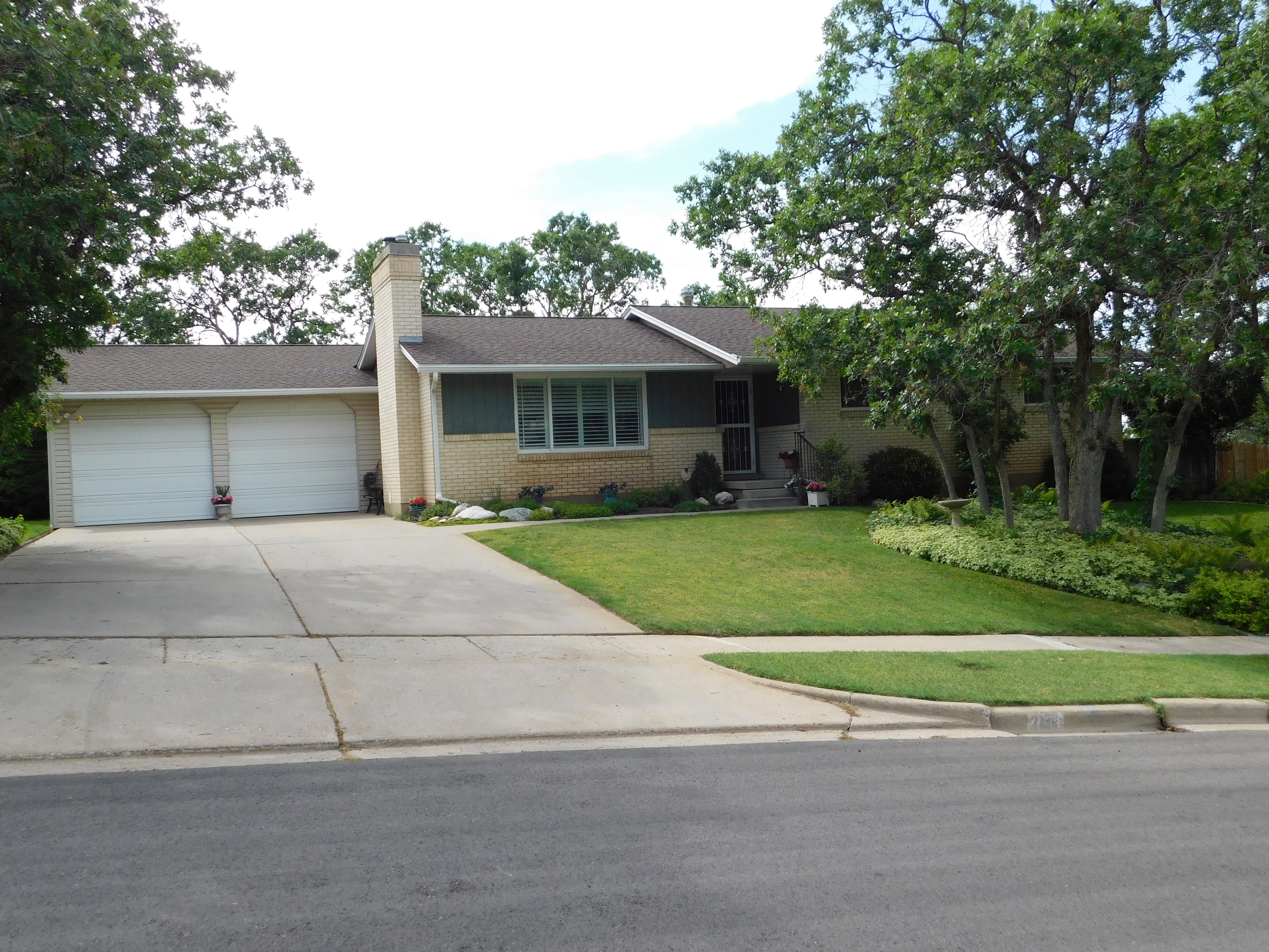 Ranch-style exterior featuring a light brick facade, dual garage doors, a shingled roof, a chimney, and mature trees