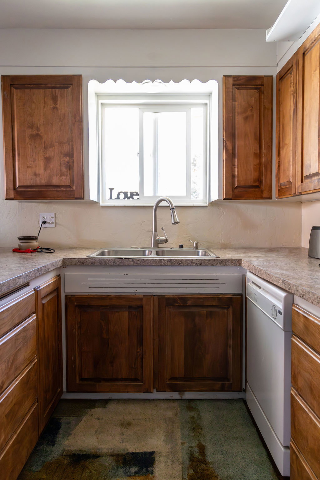 Kitchen featuring wood cabinetry, a double basin stainless steel sink with gooseneck faucet, light-toned countertops, and a built-in dishwasher