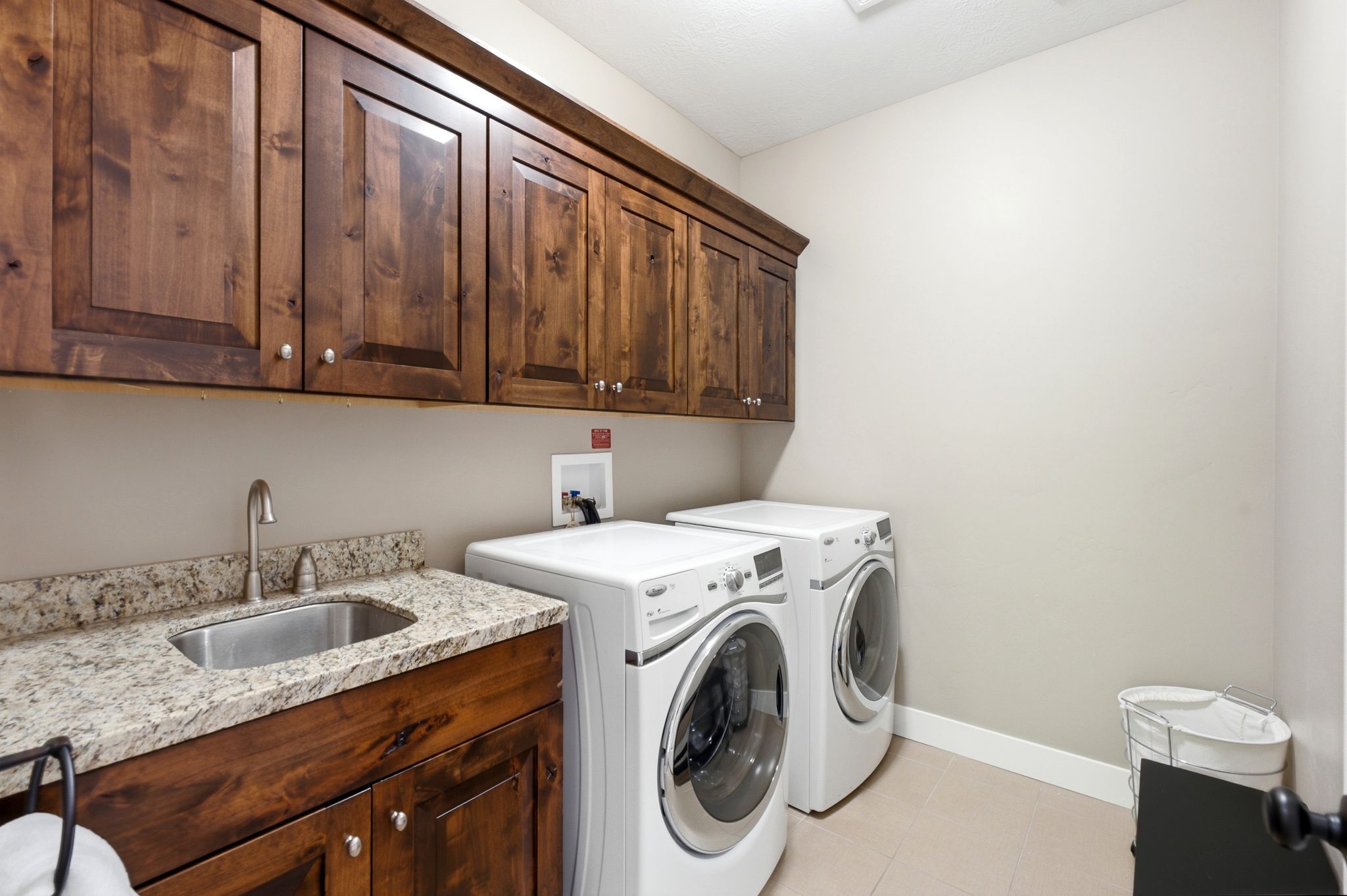 Laundry area featuring upper and lower wood cabinetry, a granite-finish countertop, a stainless steel sink with a gooseneck faucet, and light-toned floor tiling