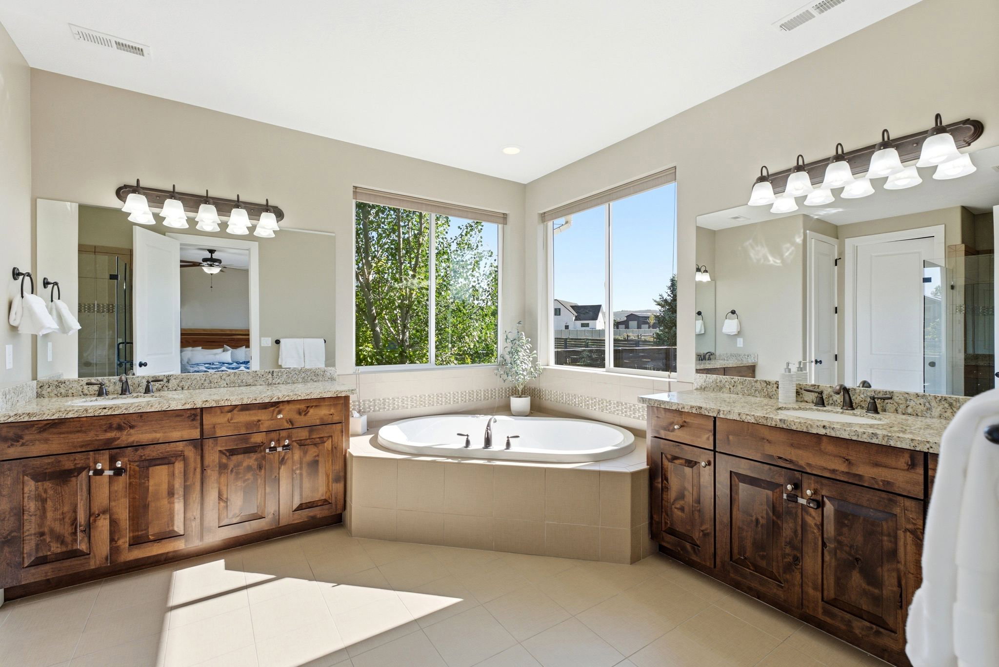 Spacious bathroom featuring an oval soaking tub, dual wood-finish vanities with light stone countertops, and large windows