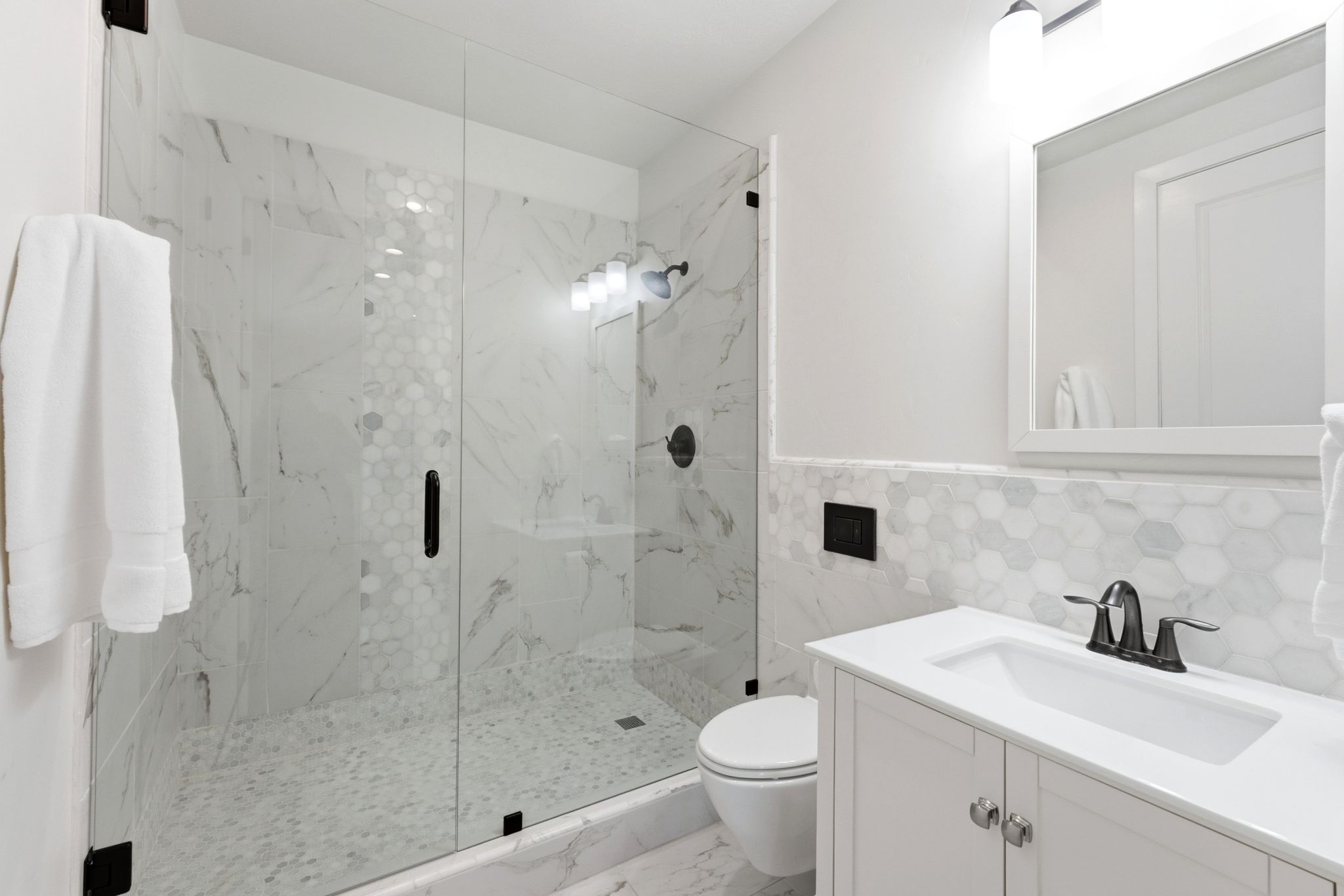 Full bathroom featuring a frameless glass shower enclosure, a white shaker-style vanity with an integrated sink, a dark bronze-finish faucet, and an illuminated mirror
