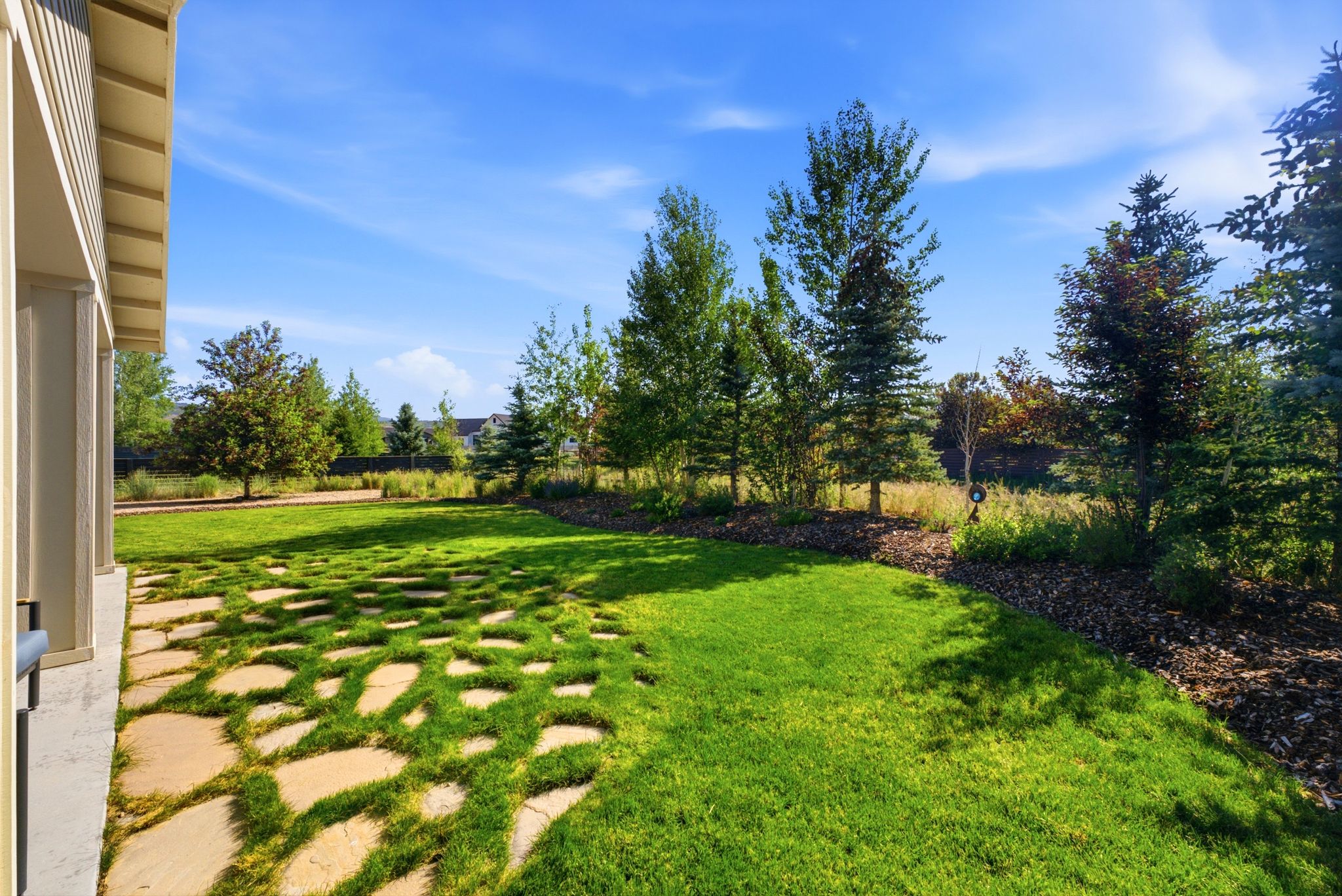 Landscaped backyard featuring a stone paver patio, manicured lawn, mature trees, and a mulched garden bed