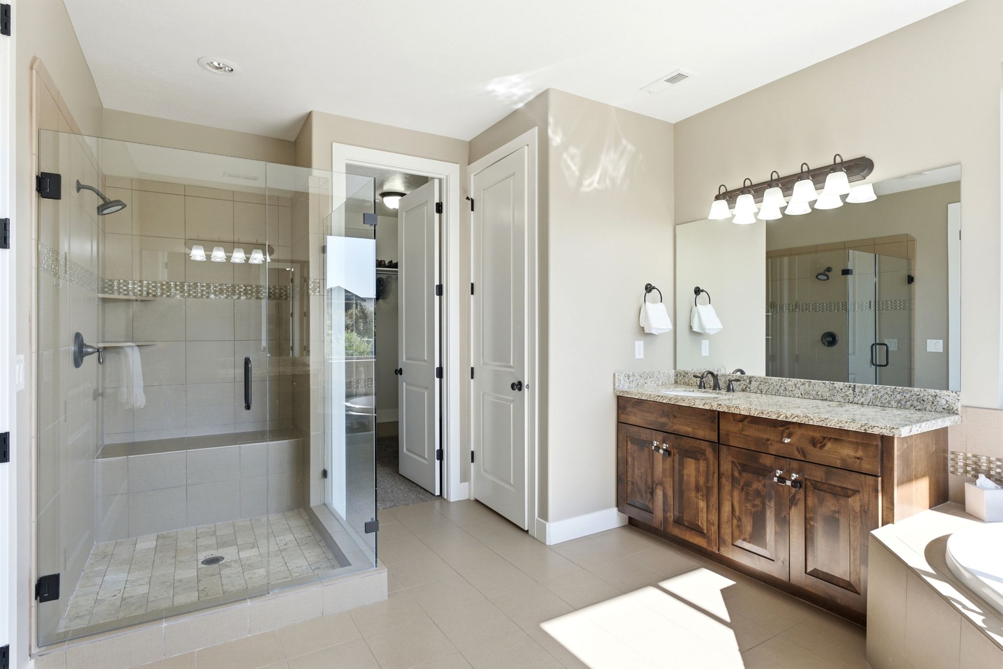 Spacious bathroom featuring a glass-enclosed shower with a built-in bench, a dual-sink vanity with a light-toned countertop, wood-finish cabinetry, and neutral tile flooring