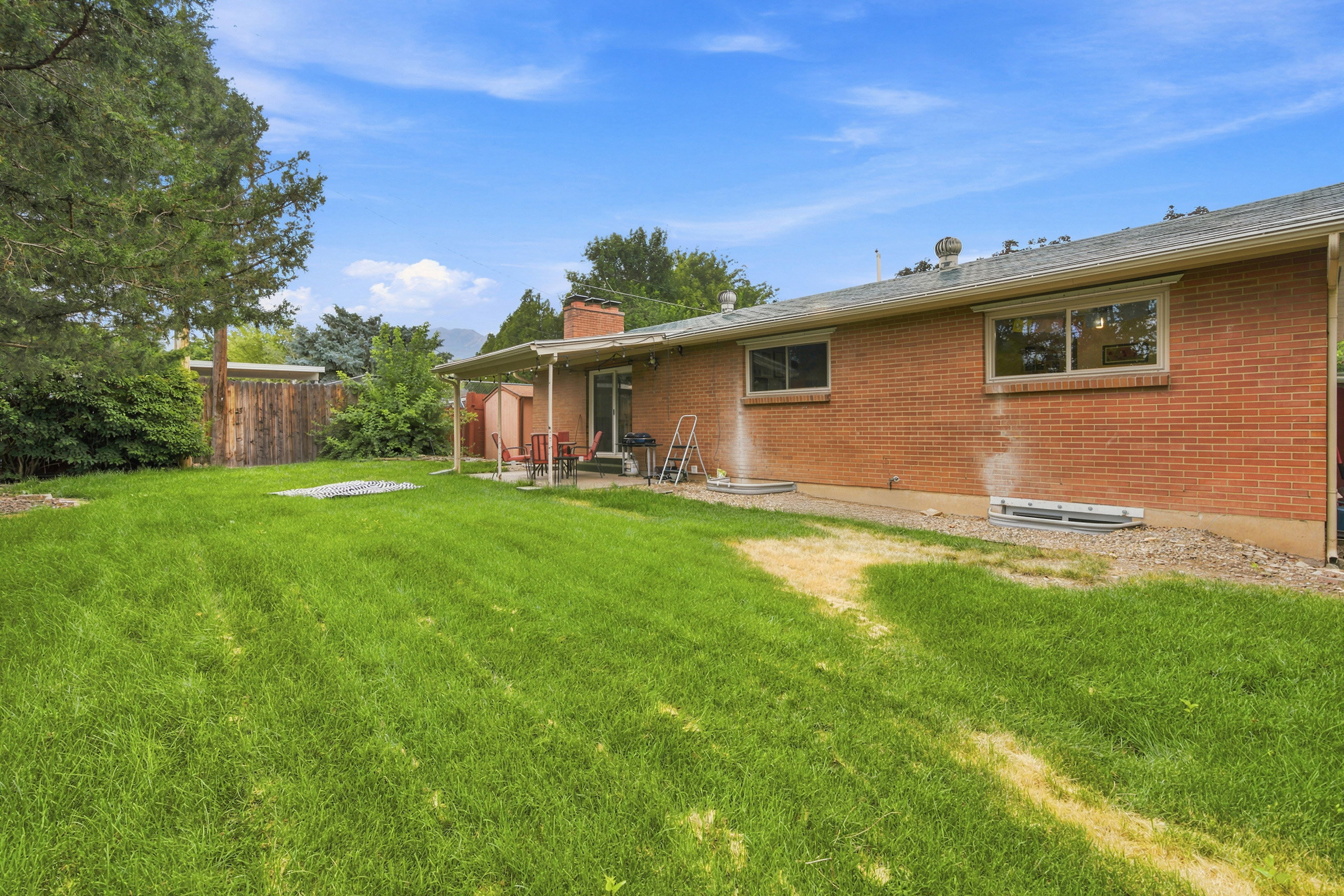Expansive backyard featuring a brick exterior with multiple windows, mature trees, and a wooden fence