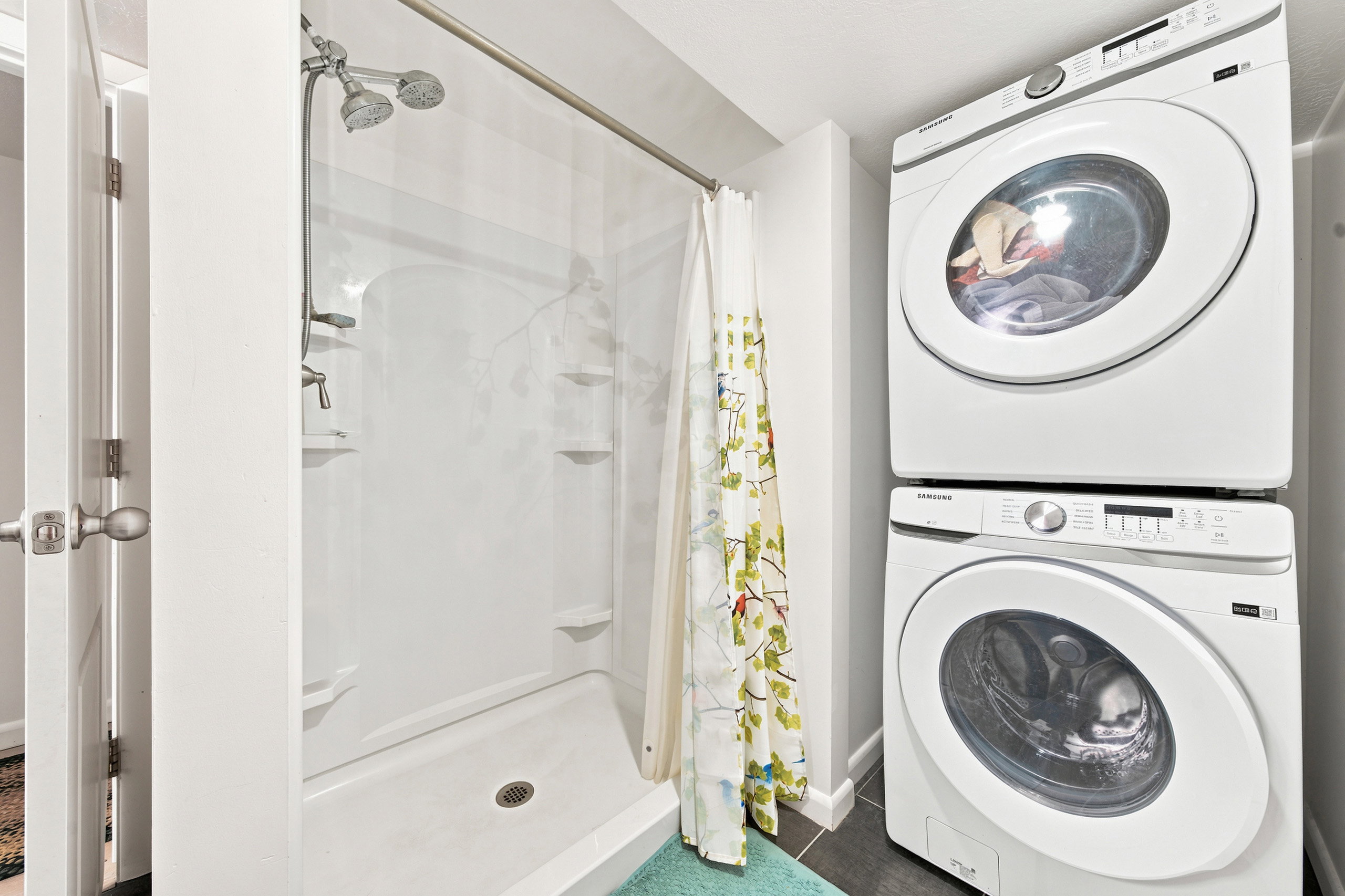 Compact bathroom featuring a shower with built-in shelving, a dual showerhead, and a stacked washer and dryer unit