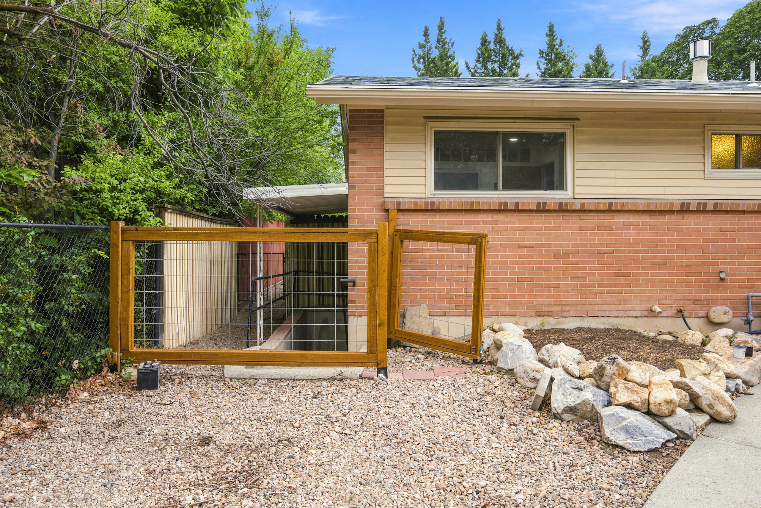 Basement apartment entrance Gravel patio area featuring a wooden gate, chain-link fence, and natural stone landscaping elements
