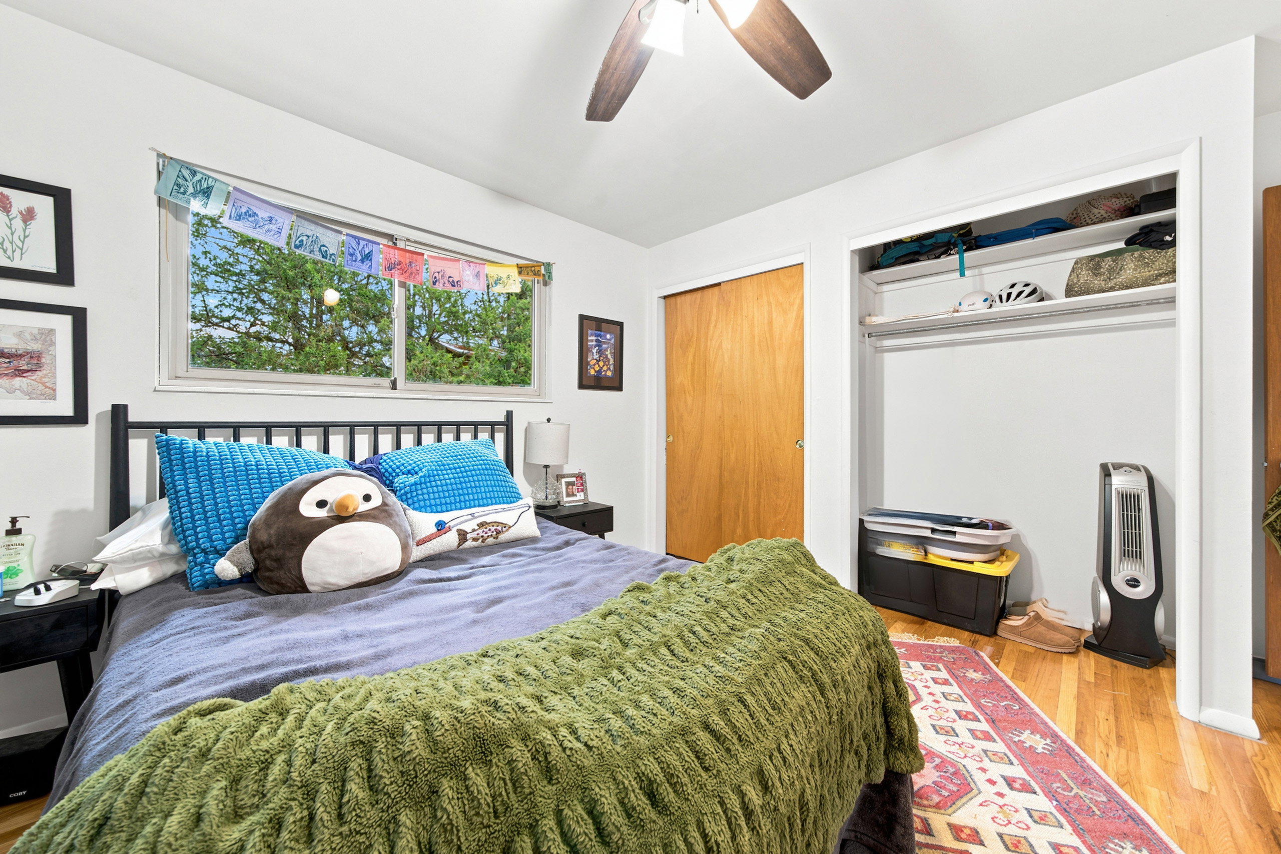 Bedroom featuring a window with exterior tree views, a ceiling fan with integrated lighting, wood-finish flooring, and a closet with shelving and hanging rod