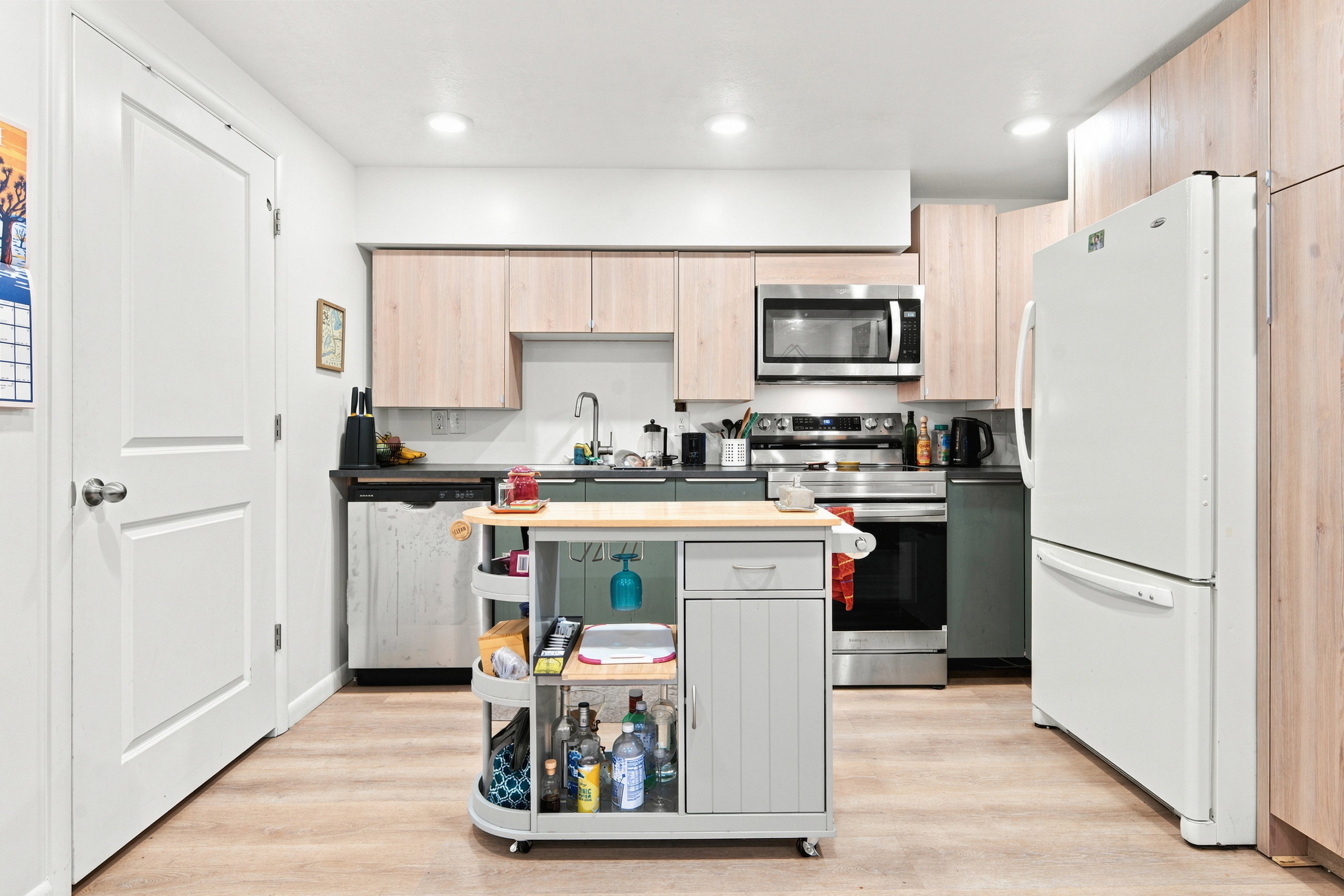 Kitchen featuring light wood-finish cabinetry, stainless steel appliances, dark countertops, and wood-finish flooring