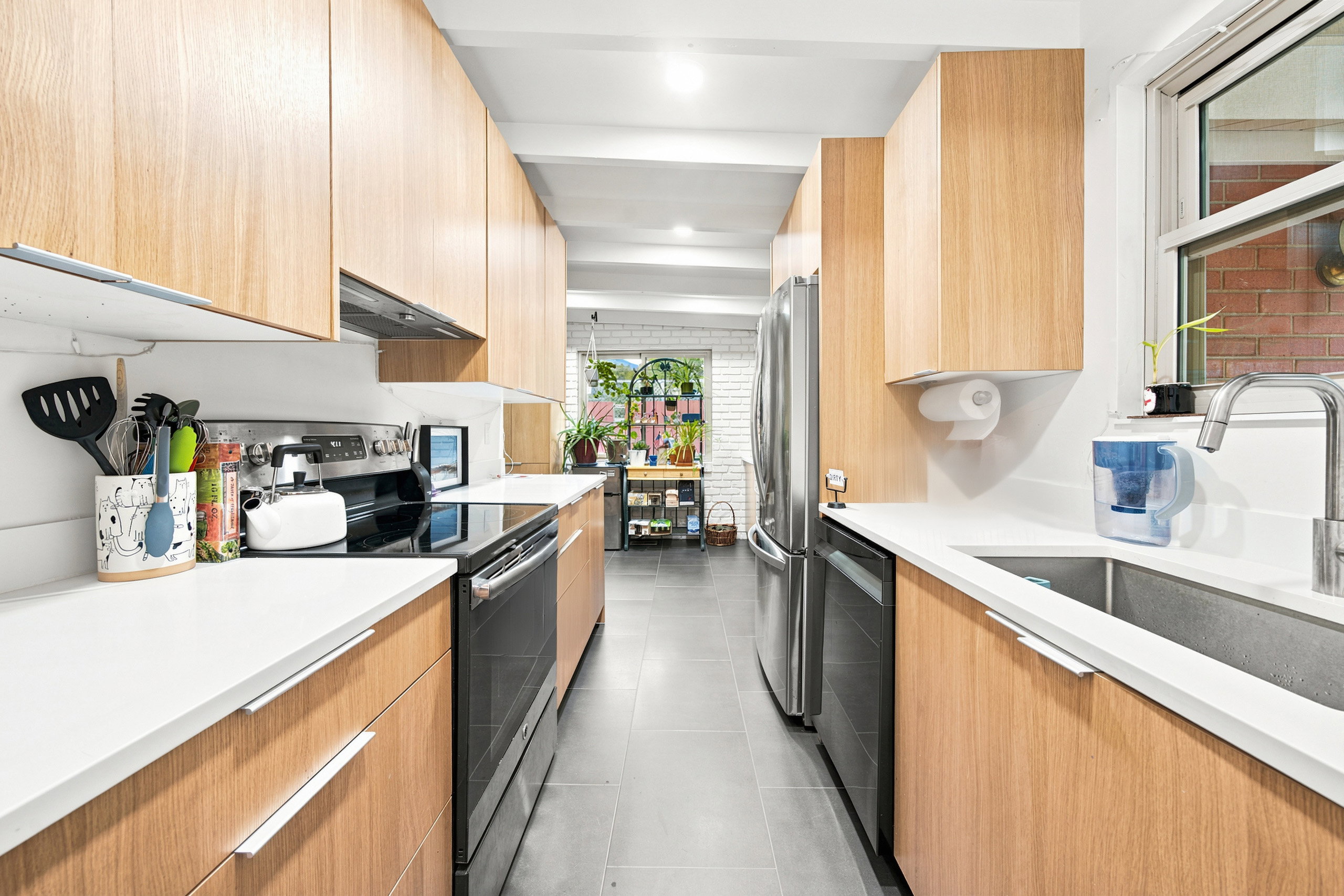 Galley kitchen featuring light wood-finish cabinetry, white solid-surface countertops, stainless steel appliances, and a stainless steel sink with a gooseneck faucet