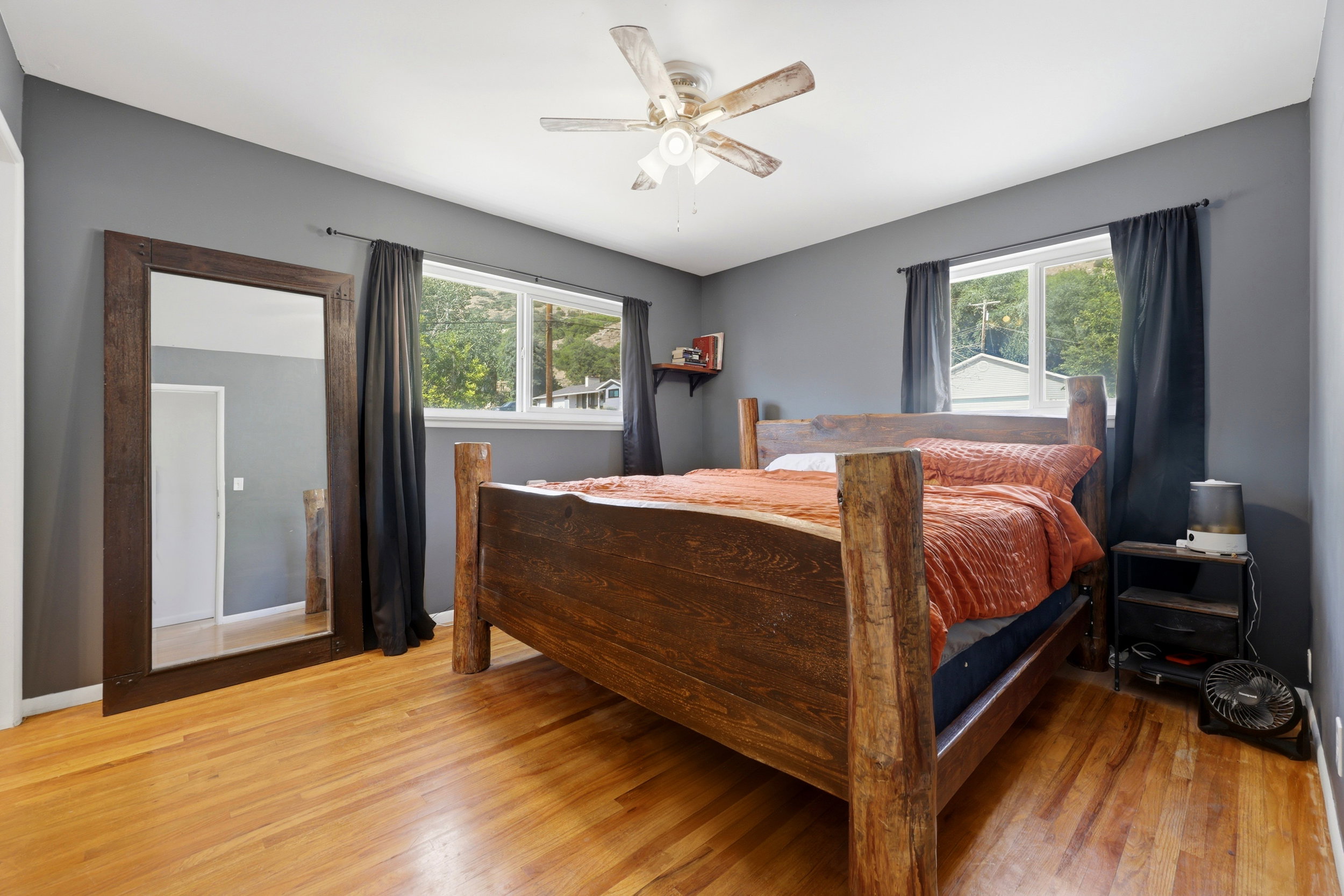 Room featuring wood-finish flooring, two large windows with dark drapery, a ceiling fan, and painted gray walls