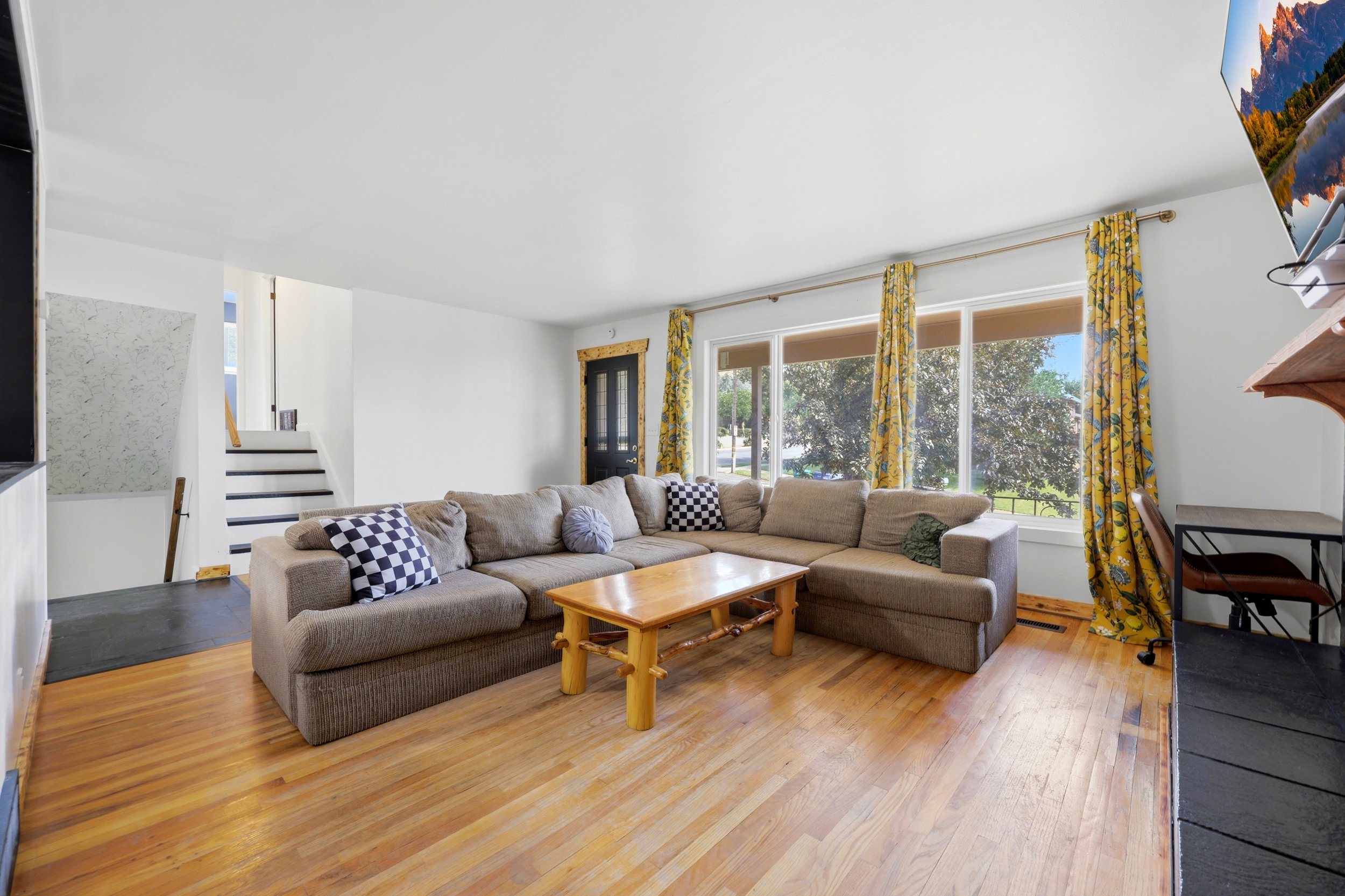 Spacious living area featuring expansive wood-finish flooring, a large window overlooking exterior greenery, white walls, and a staircase with dark treads