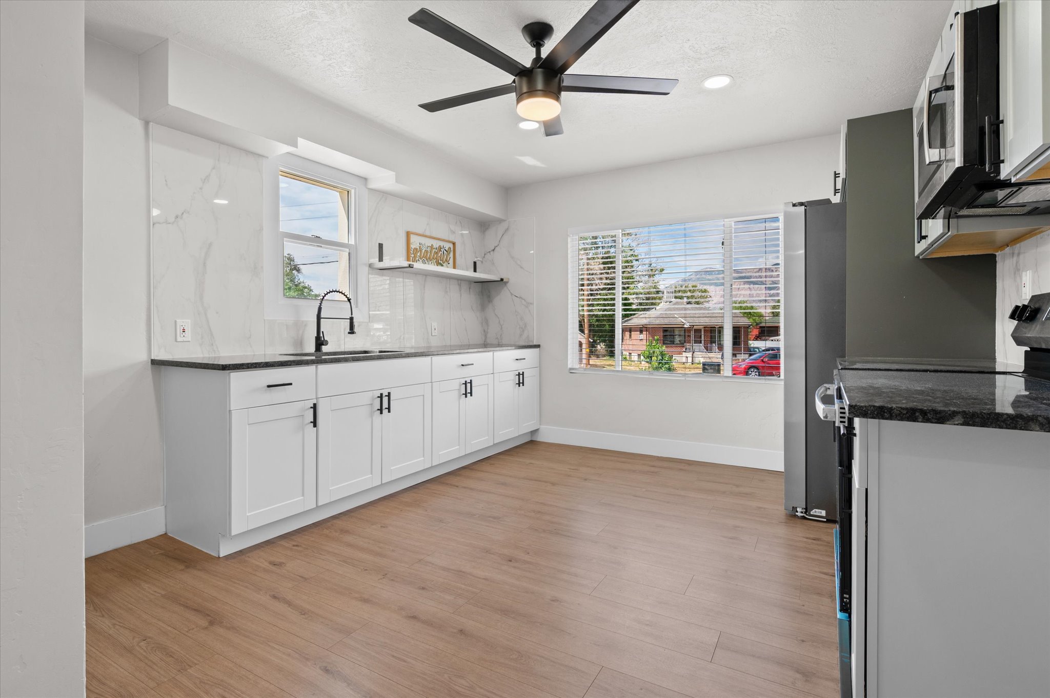 Modern kitchen featuring wood-finish flooring, white cabinetry with black hardware, dark countertops, and a white marble-look backsplash
