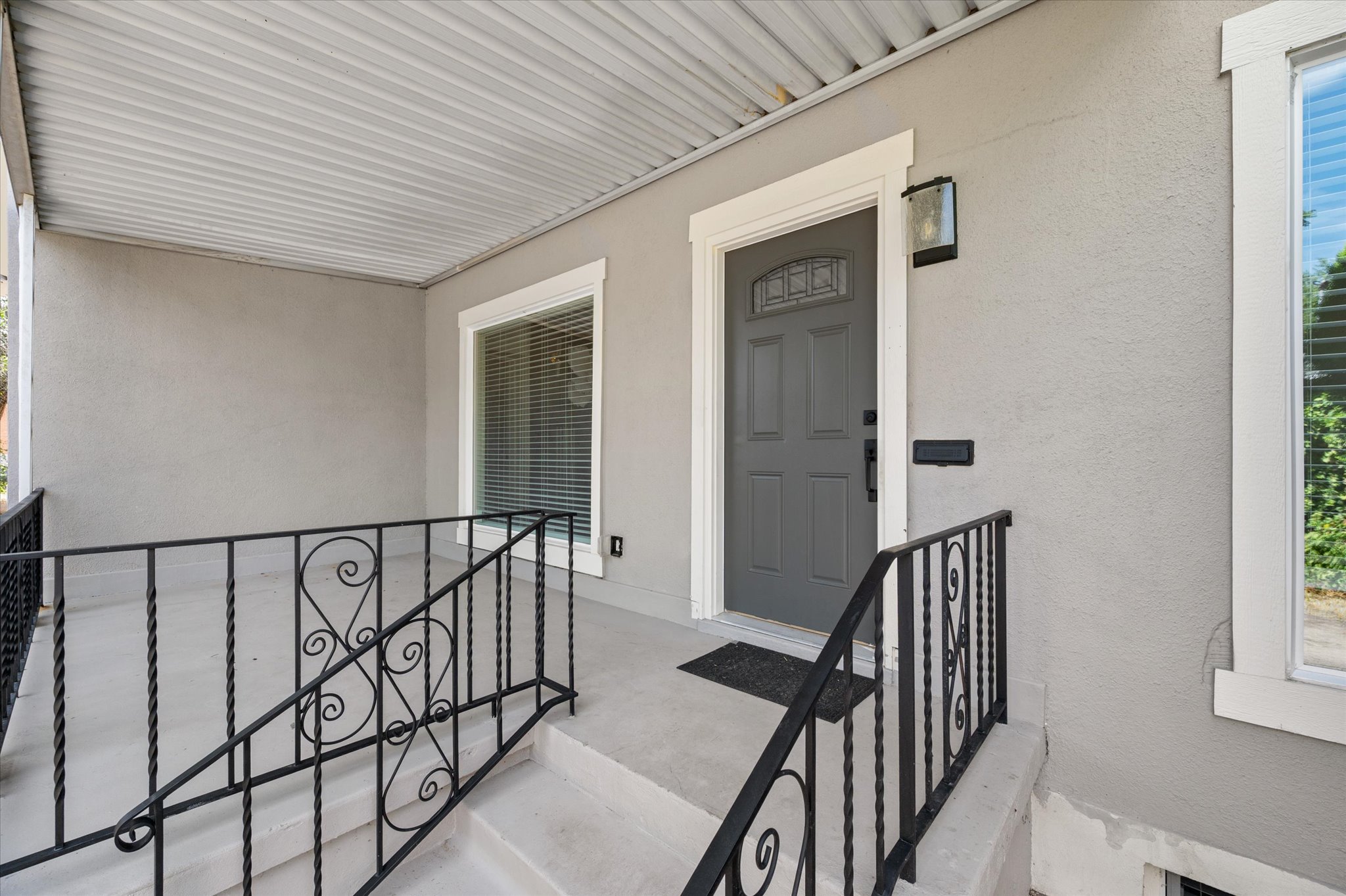 Covered entry porch featuring a paneled entry door with decorative glass, an exterior light fixture, a window with white trim, and a black wrought iron handrail