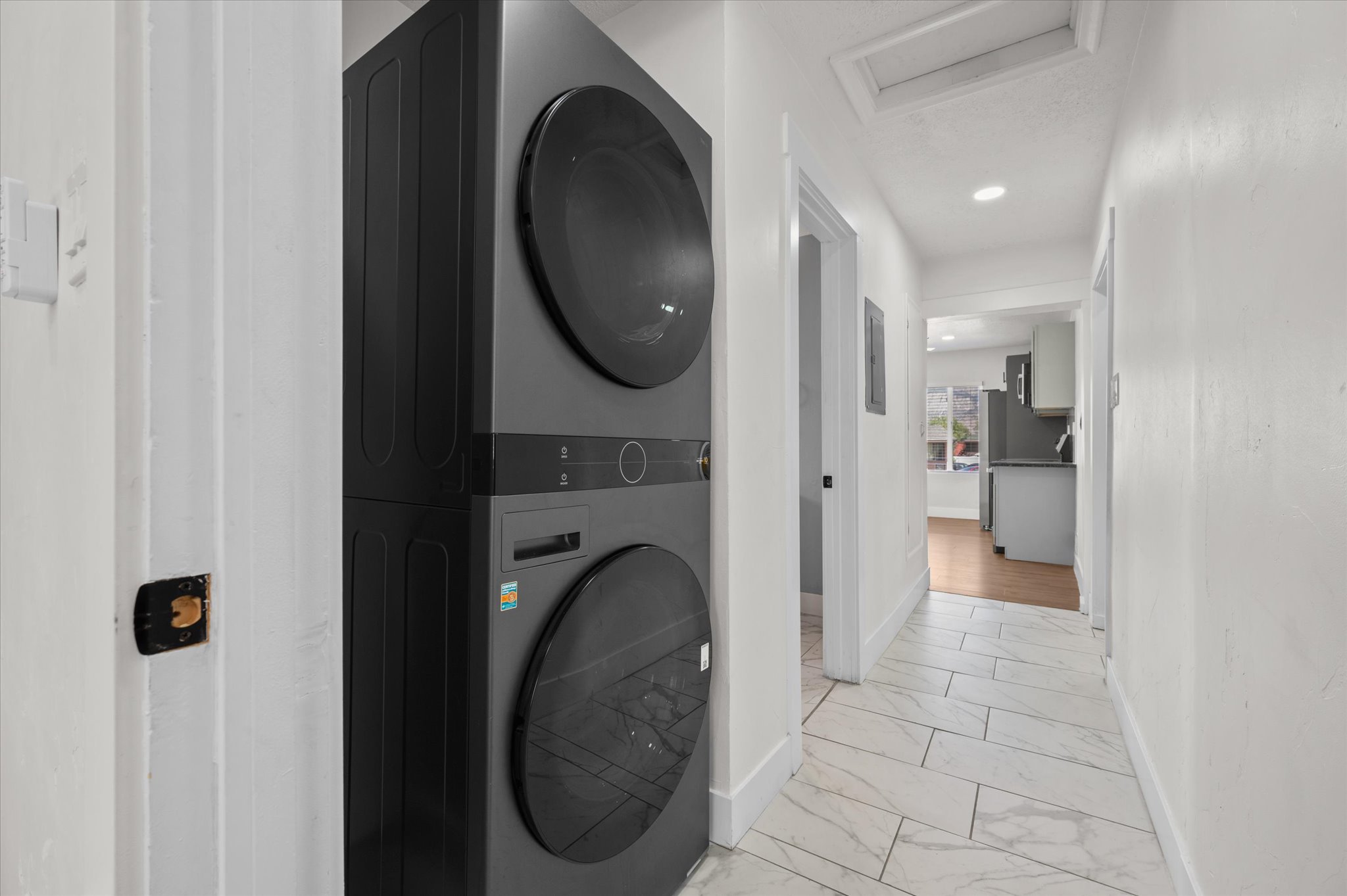 Stackable washer and dryer unit in a white-walled hallway featuring tiled flooring and recessed lighting