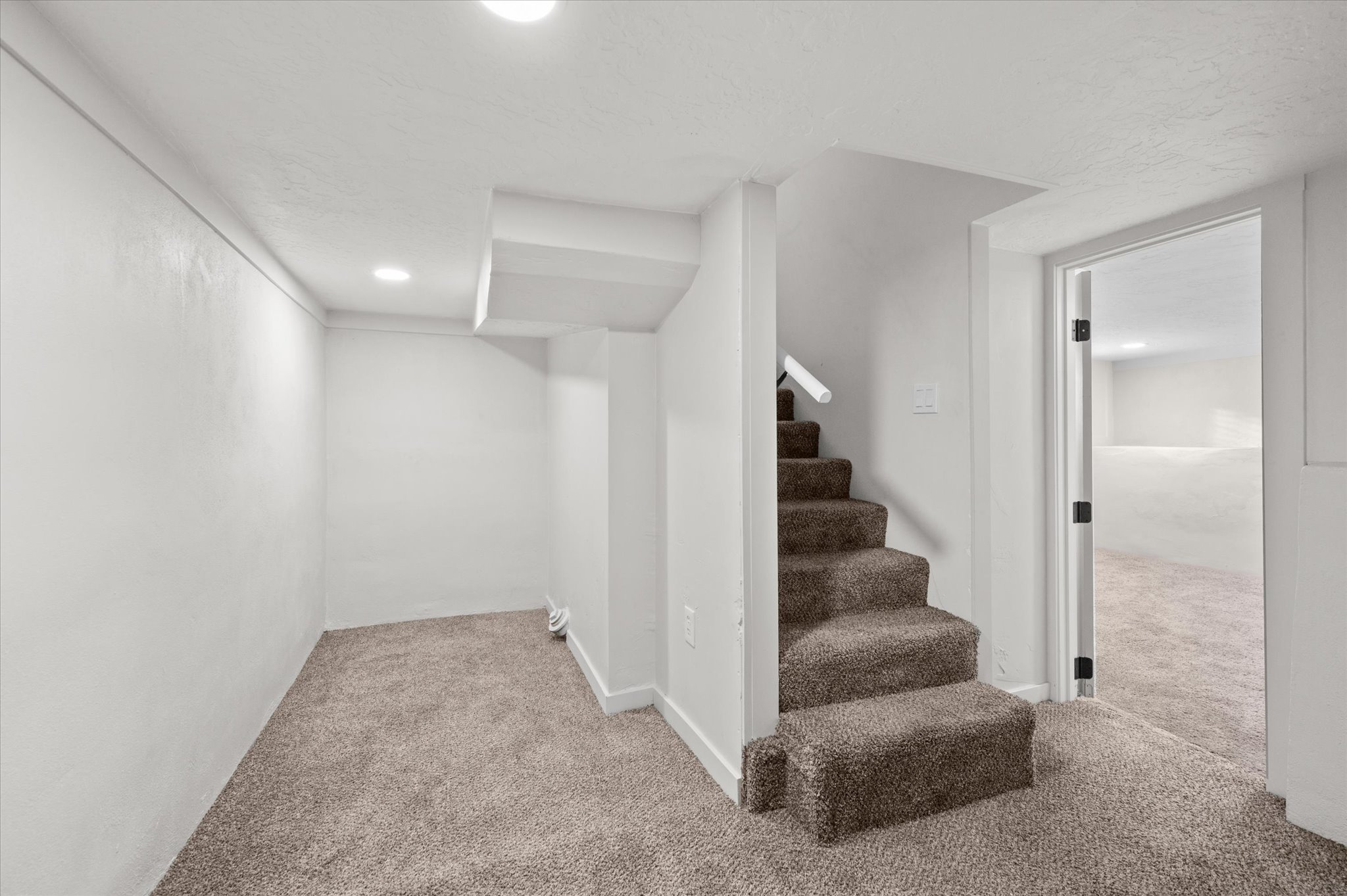 Carpeted interior space featuring a staircase with a white handrail, recessed lighting, white walls, and a doorway opening to an additional room