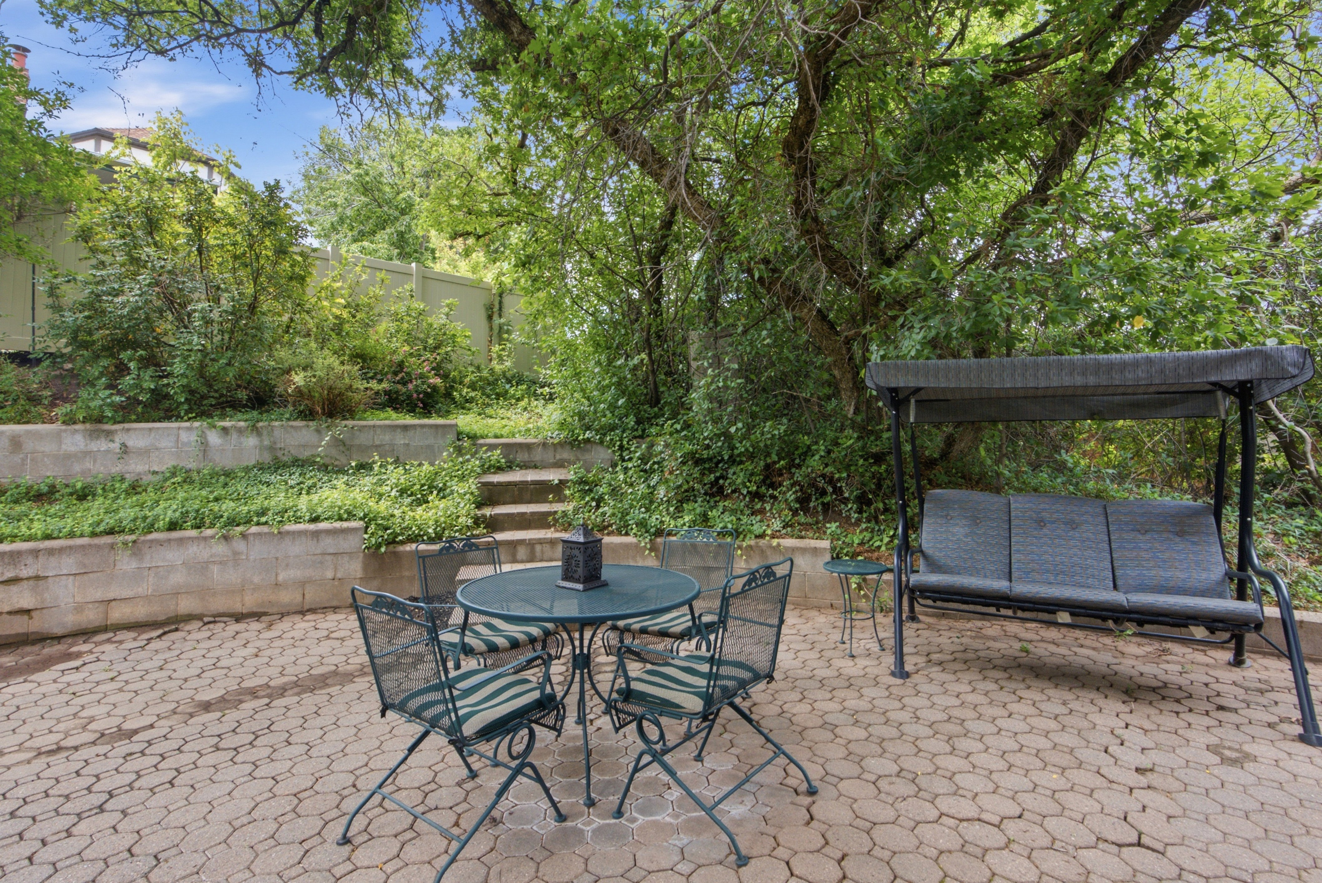 Paved patio featuring a geometric paver design, surrounded by mature trees and layered garden beds with concrete retaining walls