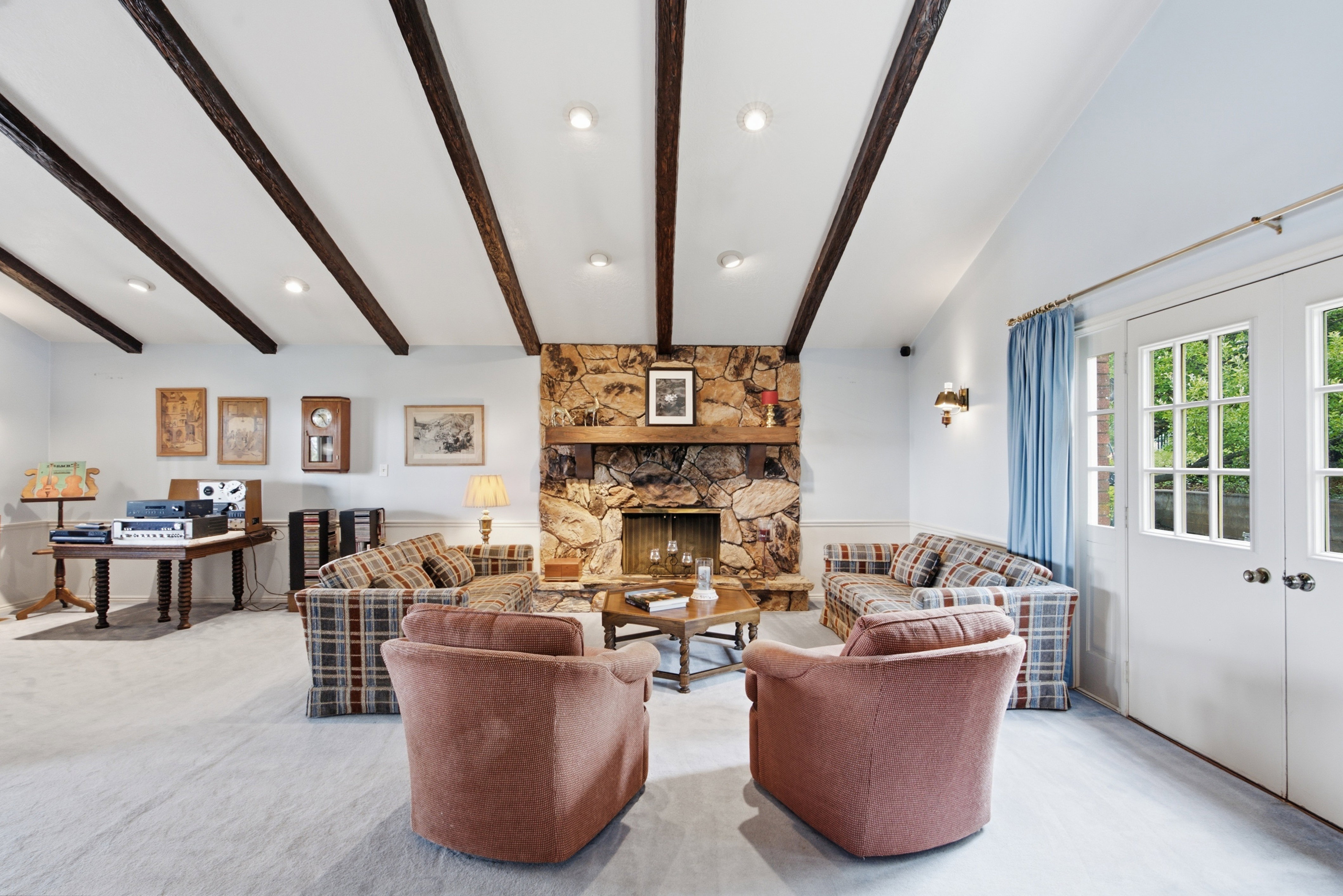 Spacious living area featuring a stone-clad fireplace with a wooden mantel, exposed ceiling beams, recessed lighting, and light-toned carpeting