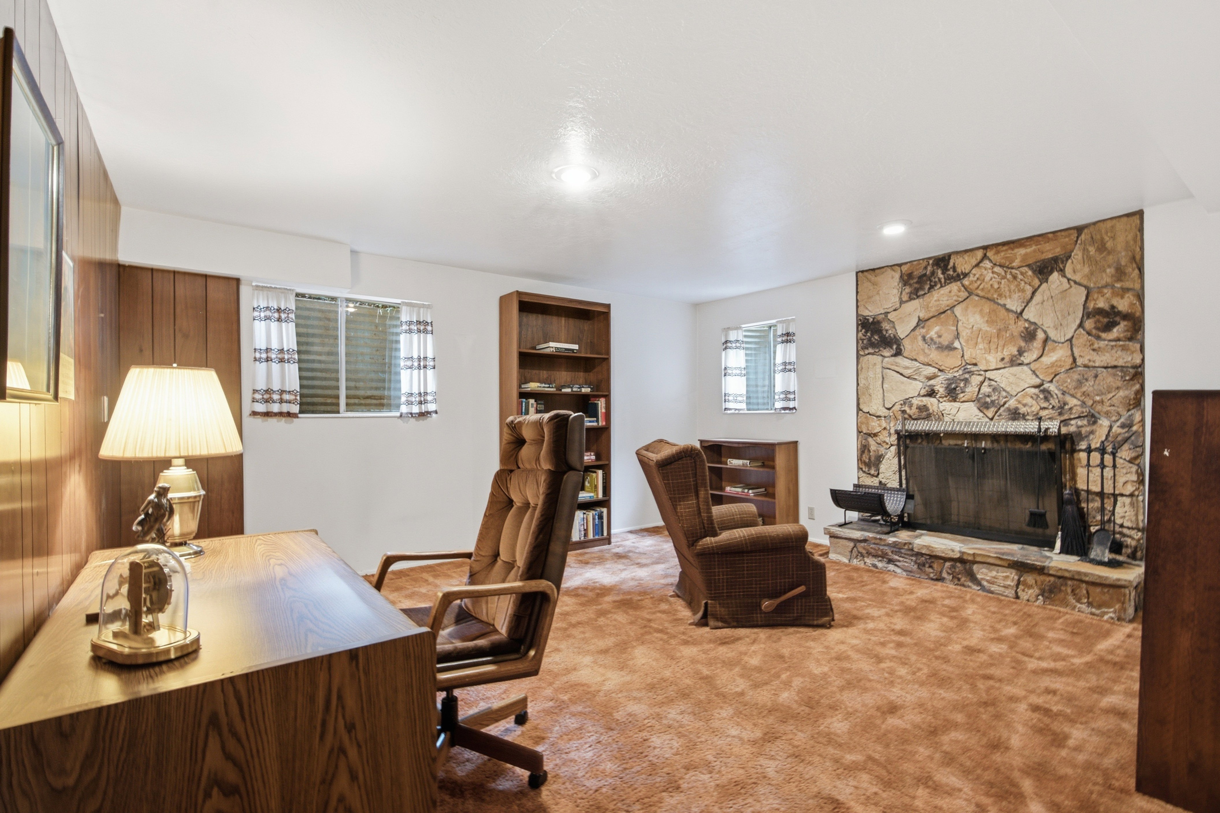 Spacious room featuring a stone-clad fireplace with a hearth, wood paneling, wood-finish shelving, and two windows with blinds