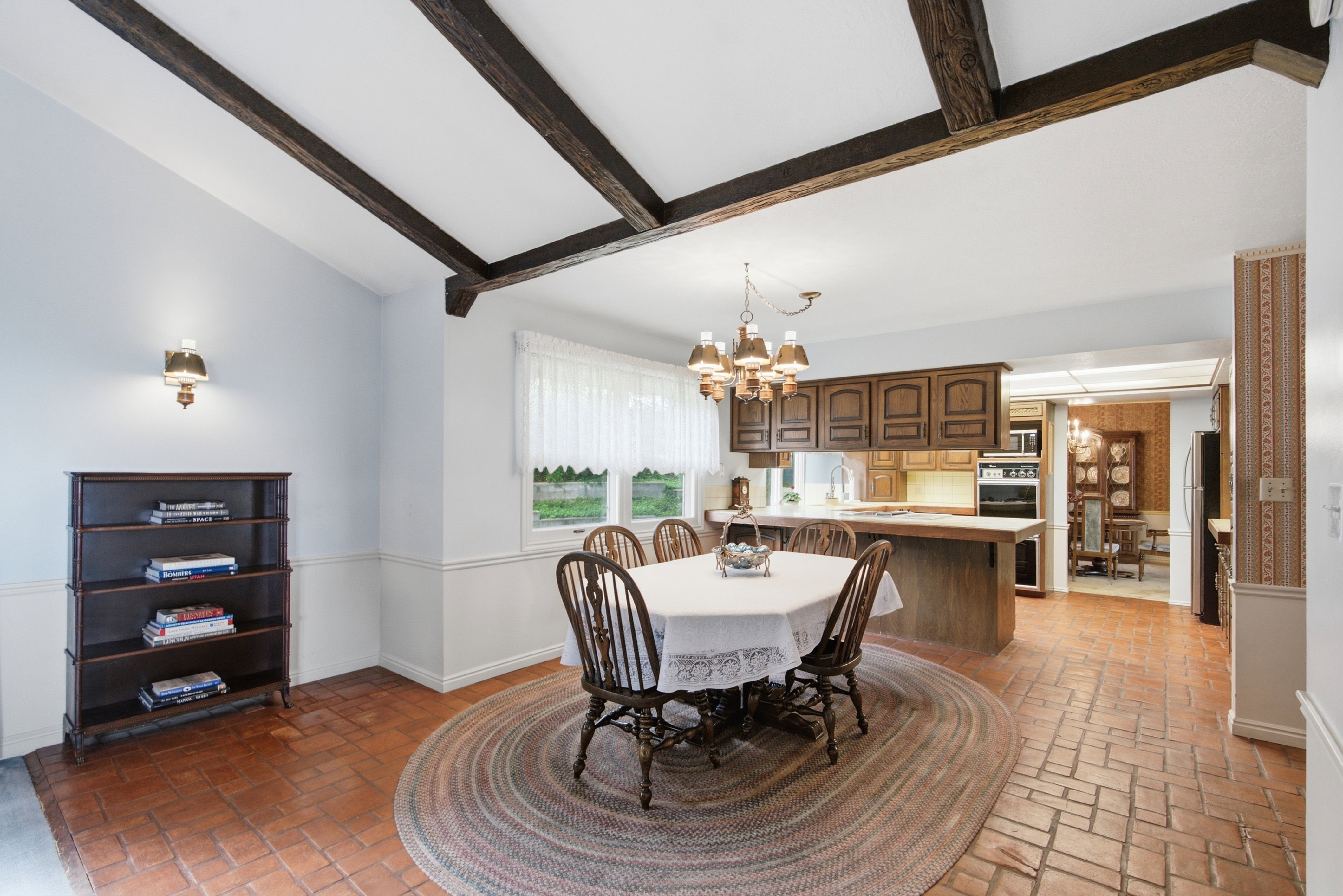 Vaulted ceiling with exposed wood beams, brick-finish flooring, wood cabinetry, built-in wall oven, and a chandelier fixture