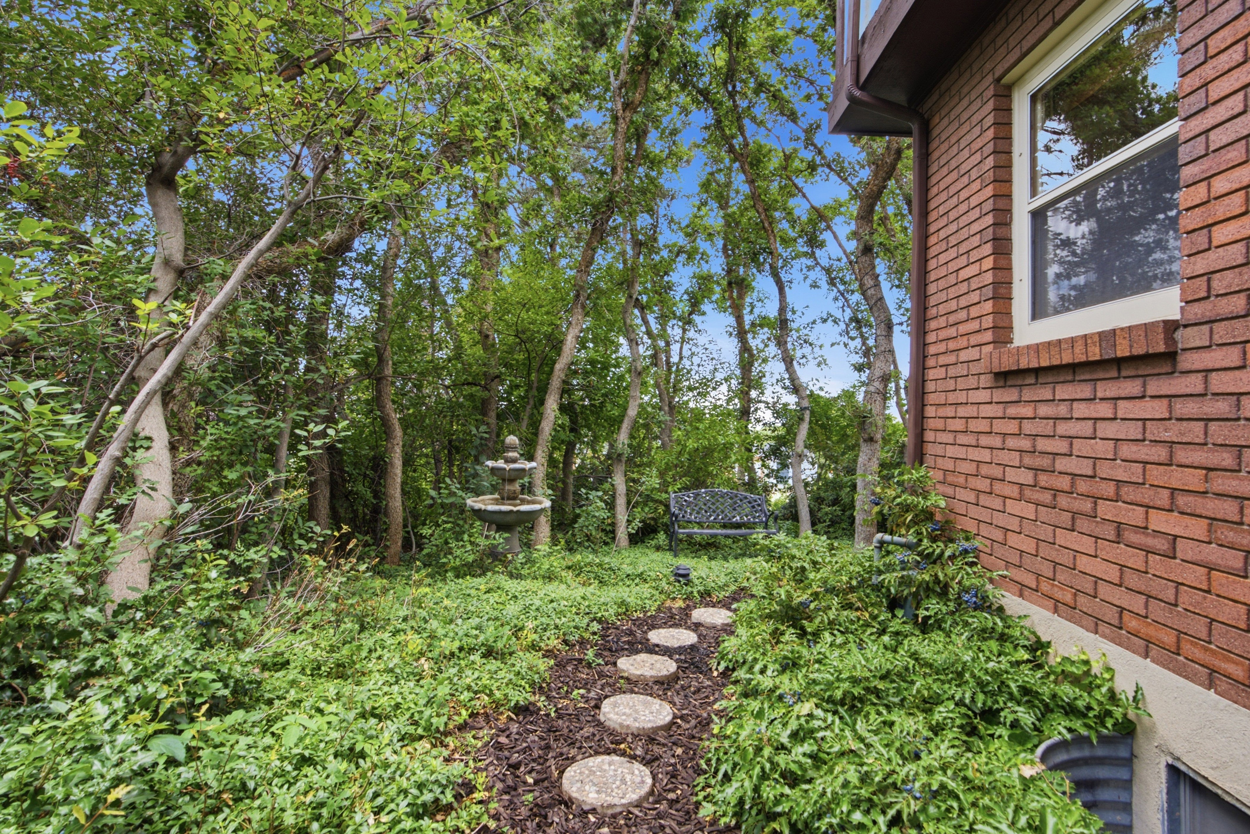 Established landscaping featuring mature trees, a multi-tiered fountain, and a stepping stone path
