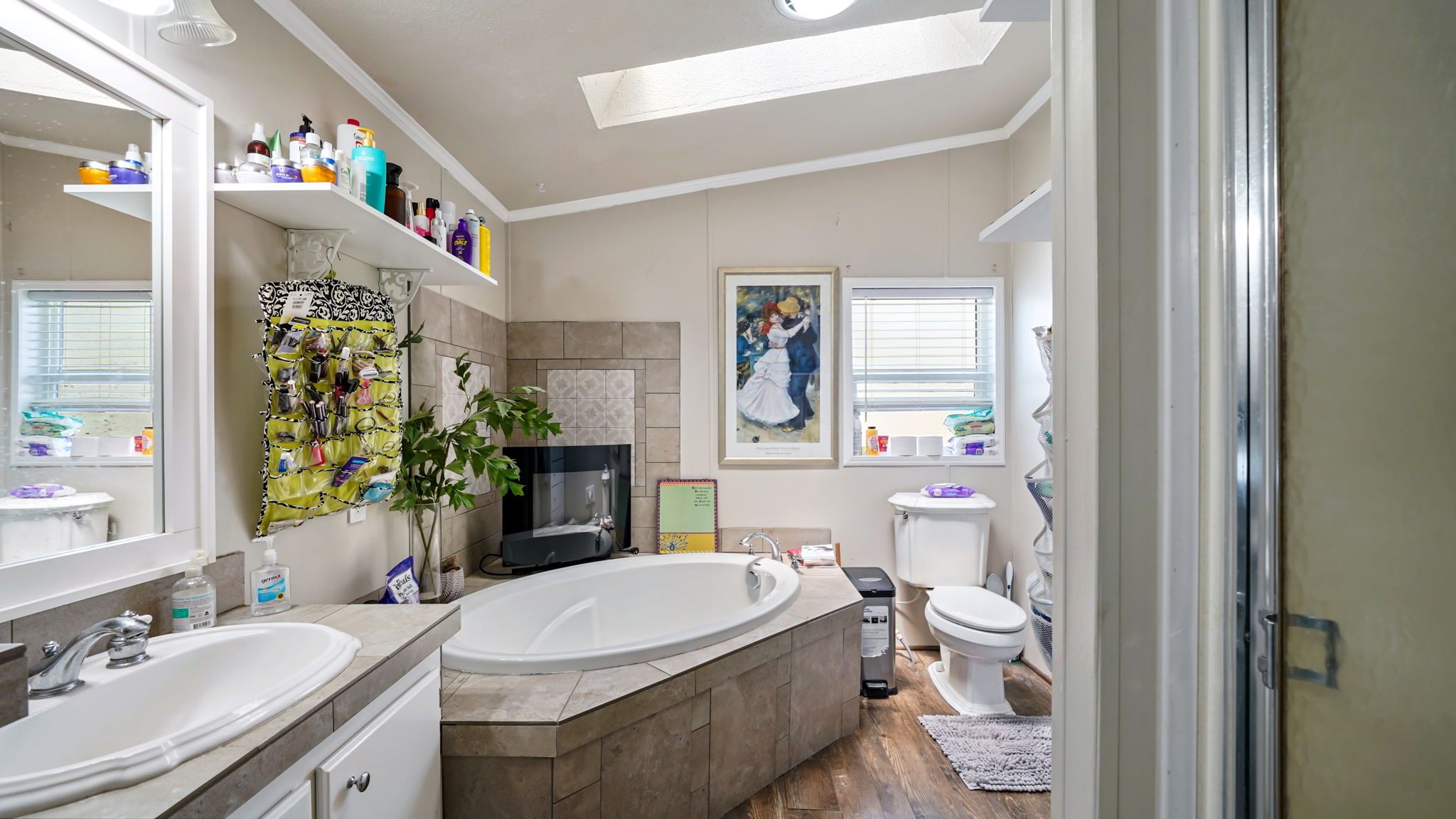 Bathroom featuring a built-in soaking tub with a tiled surround, a vanity with dual sinks and a framed mirror, wood-finish flooring, a skylight, and a separate shower enclosure