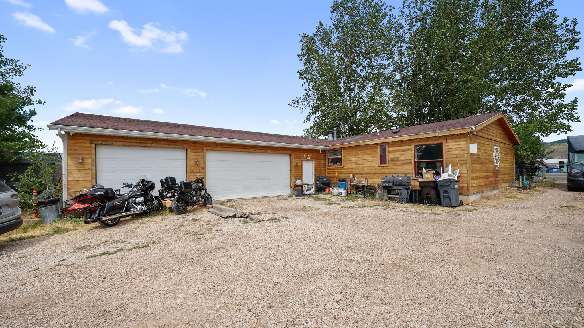 Wood-sided structure featuring a two-car garage with white doors, a brown shingle roof, and exterior lighting fixtures