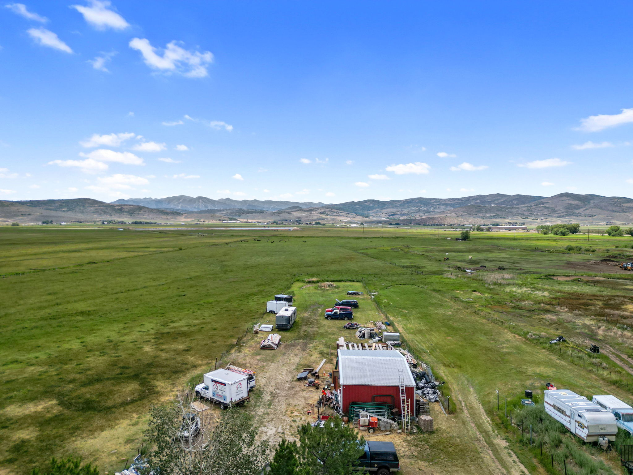 Expansive rural property featuring a red barn with a metal roof, surrounded by green pastureland, a gravel drive, and distant mountains under a blue sky