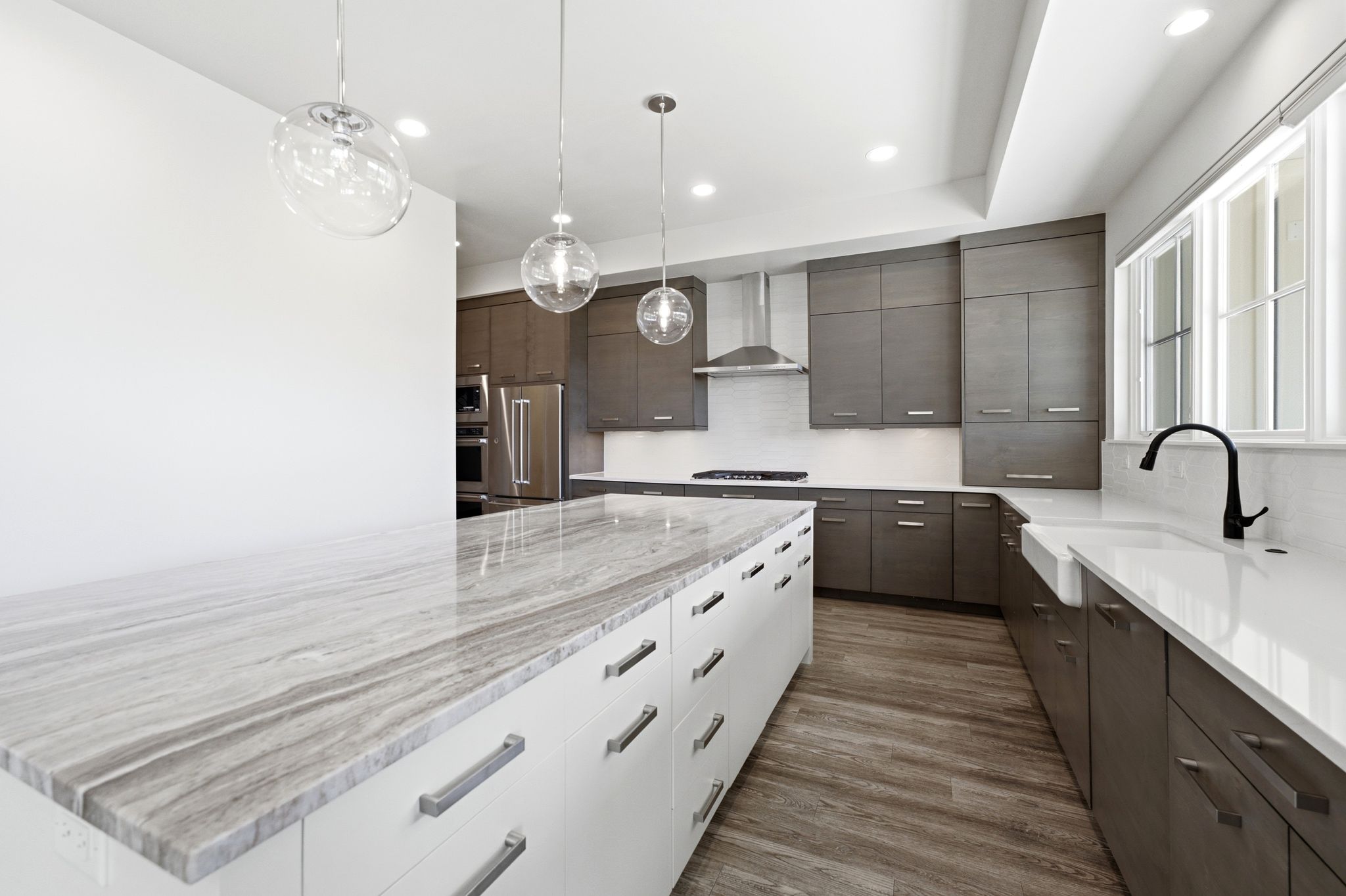 Contemporary kitchen featuring a stone-top island with extensive drawer storage, wood-finish flooring, dark-toned cabinetry, and a white farmhouse sink with a matte black faucet