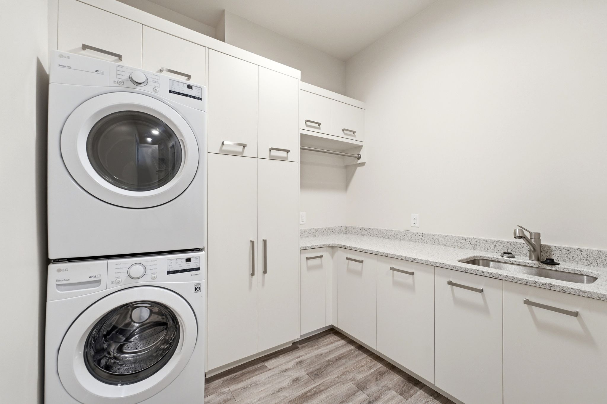 Dedicated laundry area featuring stacked front-loading appliances, extensive white cabinetry, a built-in sink, light speckled countertops, and wood-finish flooring