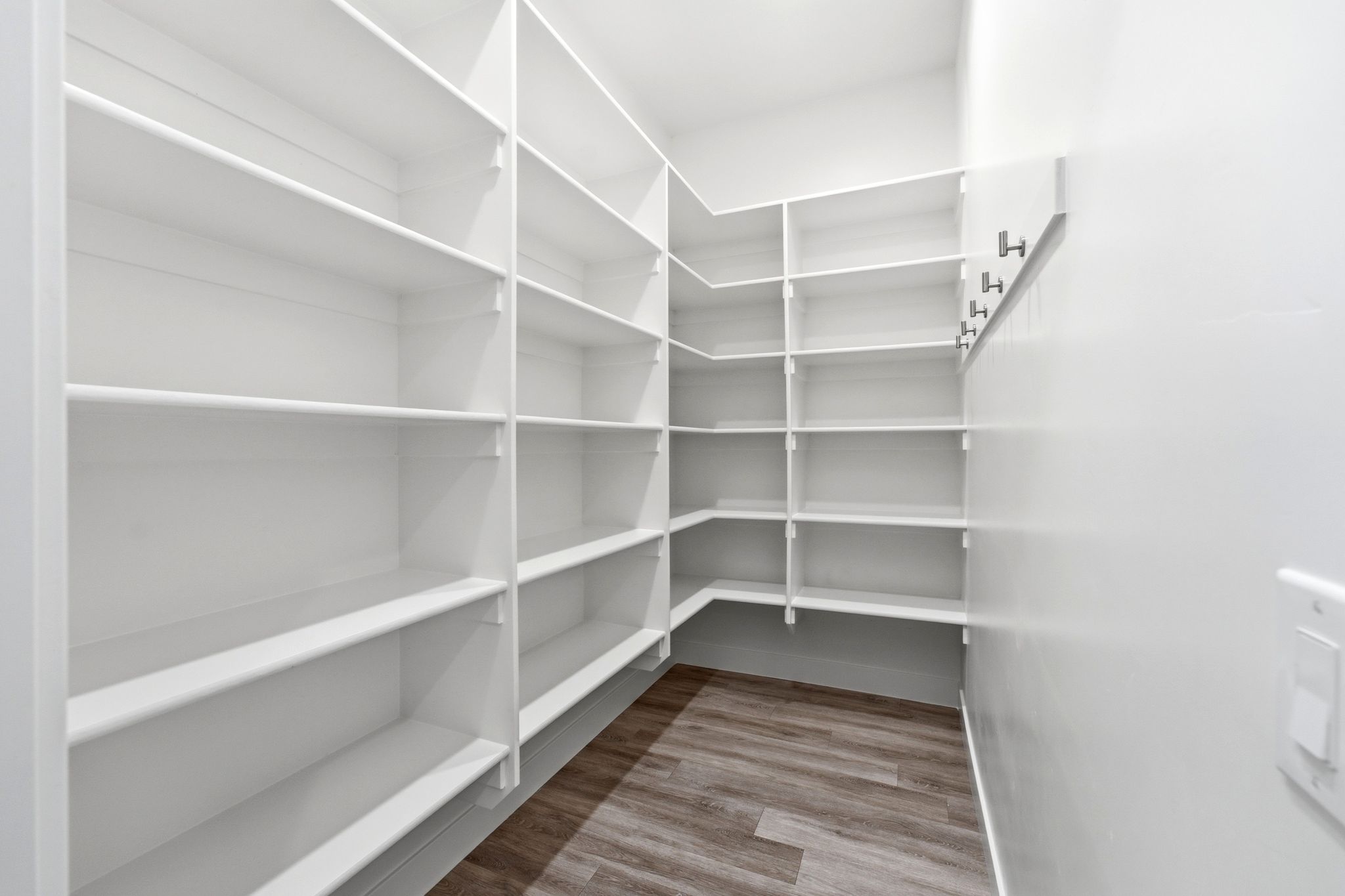 Spacious walk-in pantry featuring white shelving, light wood-finish flooring, and wall-mounted hooks