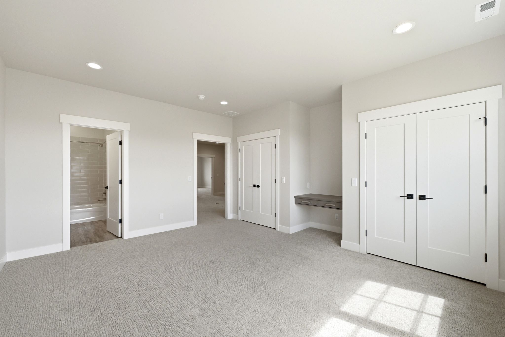Spacious room featuring light gray carpeting, recessed lighting, white trim, a built-in floating desk, and multiple white panel doors with matte black hardware