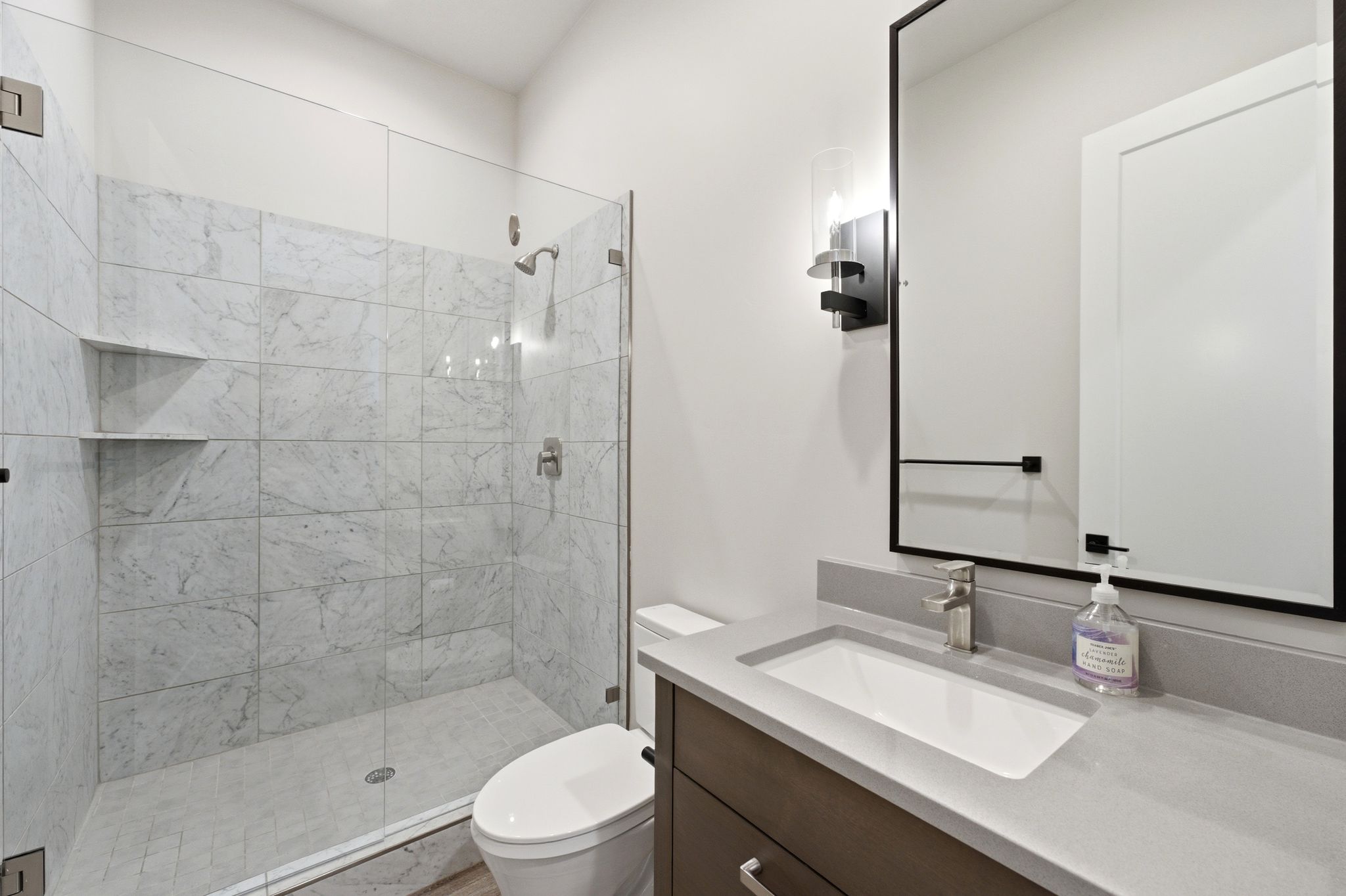 Bathroom featuring a glass-enclosed shower with tiled walls and built-in shelving, a contemporary vanity with an integrated sink and light grey countertop, and a sleek mirror with a matte black frame