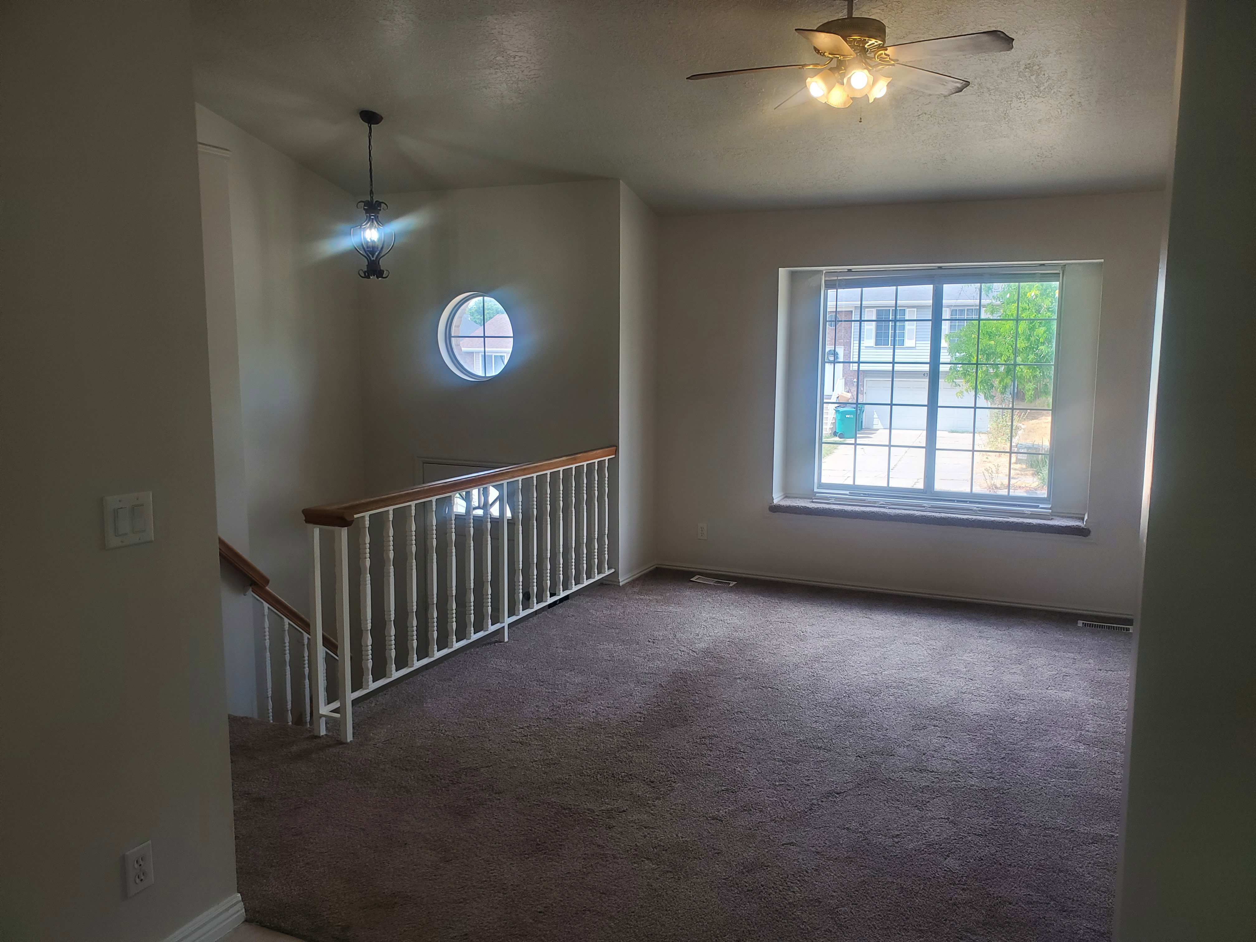 Carpeted interior space featuring a large picture window, a ceiling fan with integrated lighting, a decorative pendant light fixture, a round accent window, and a staircase with a white balustrade and wood-finish handrail