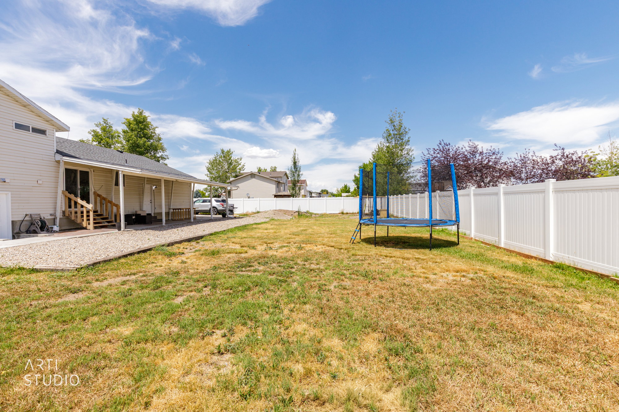 Expansive backyard featuring a white vinyl privacy fence, a covered patio with a gravel path, and wood exterior stairs
