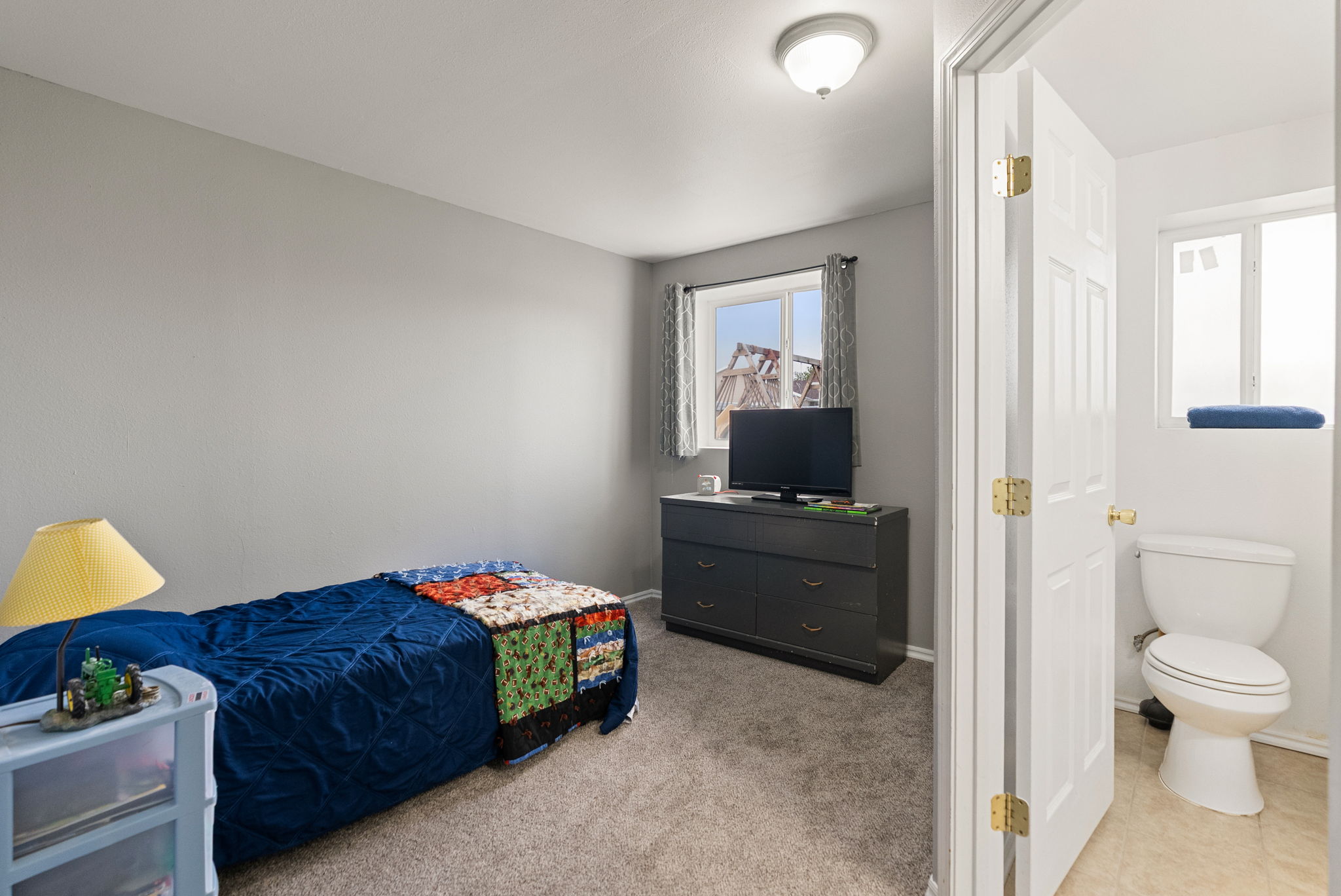 Carpeted bedroom featuring a window, built-in overhead lighting, and an entryway to an attached bathroom with a toilet and privacy window