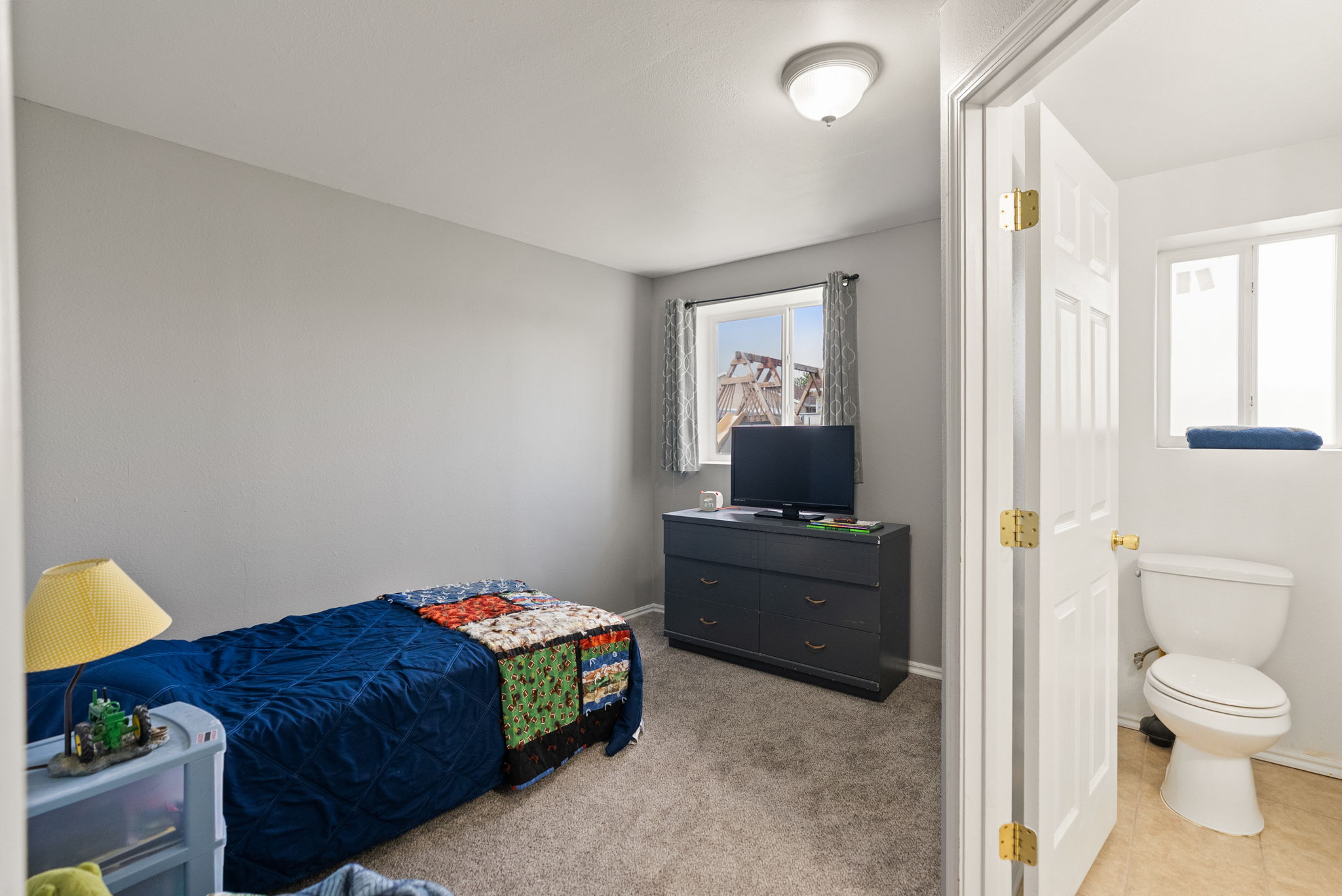 Carpeted bedroom featuring a window with blinds, a ceiling-mounted light fixture, and a nearby bathroom with a toilet and window